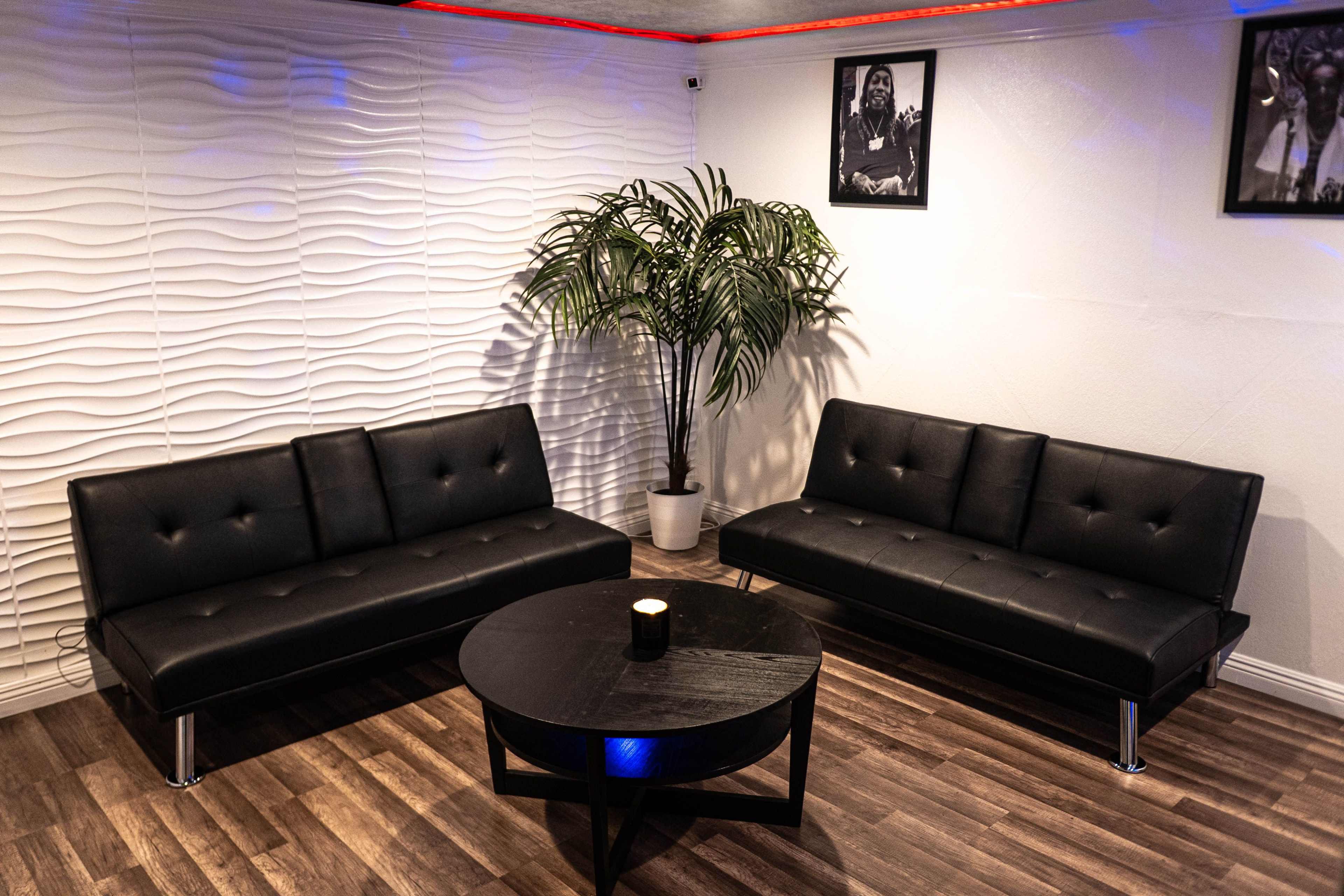 The image shows a modern lounge area featuring two black sofas, a round black table, and a potted plant, with textured white walls and framed photographs in the background.