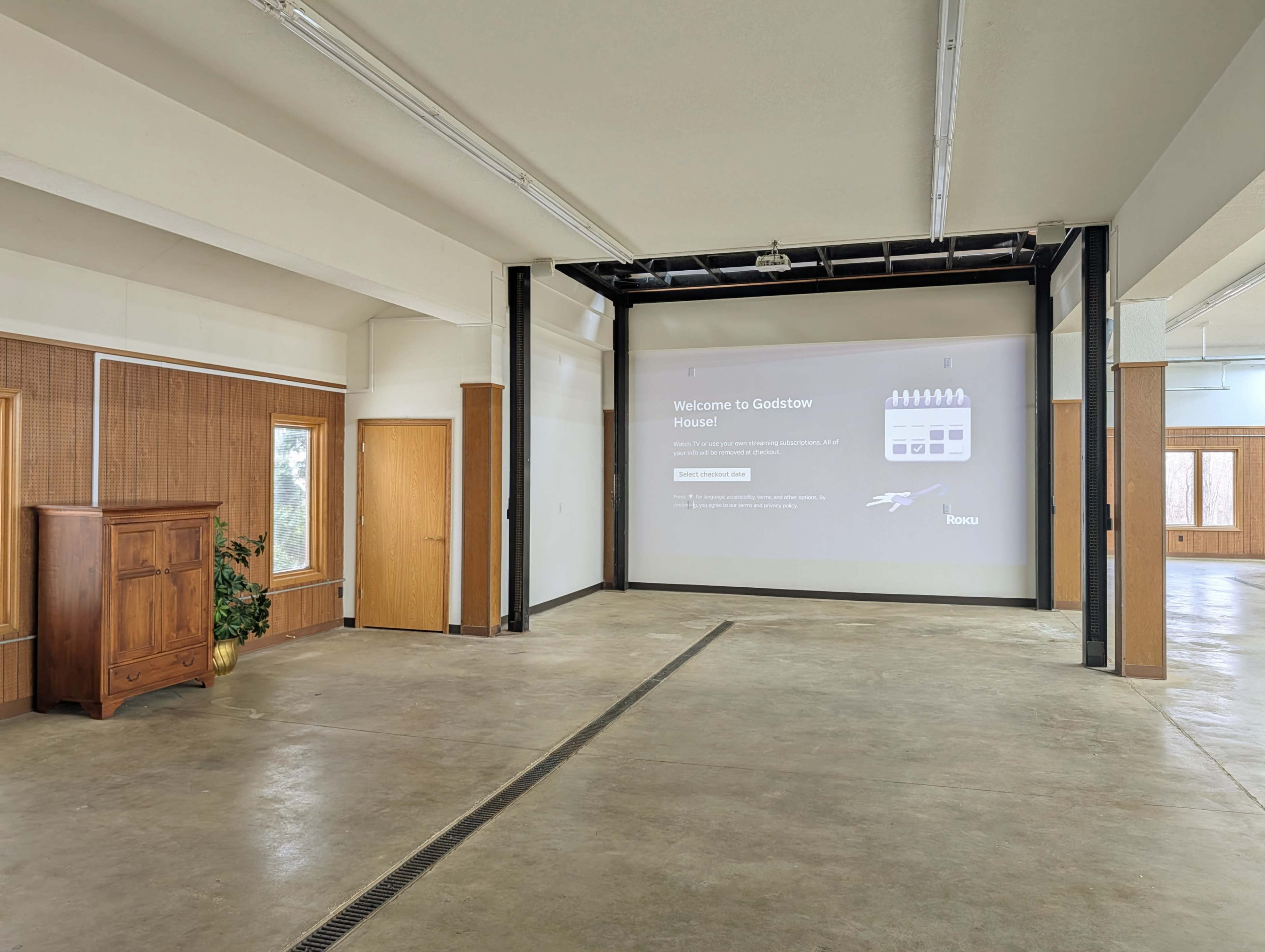 The image shows an empty interior room featuring a large projection screen on one wall and wooden doors and furniture on the other walls.