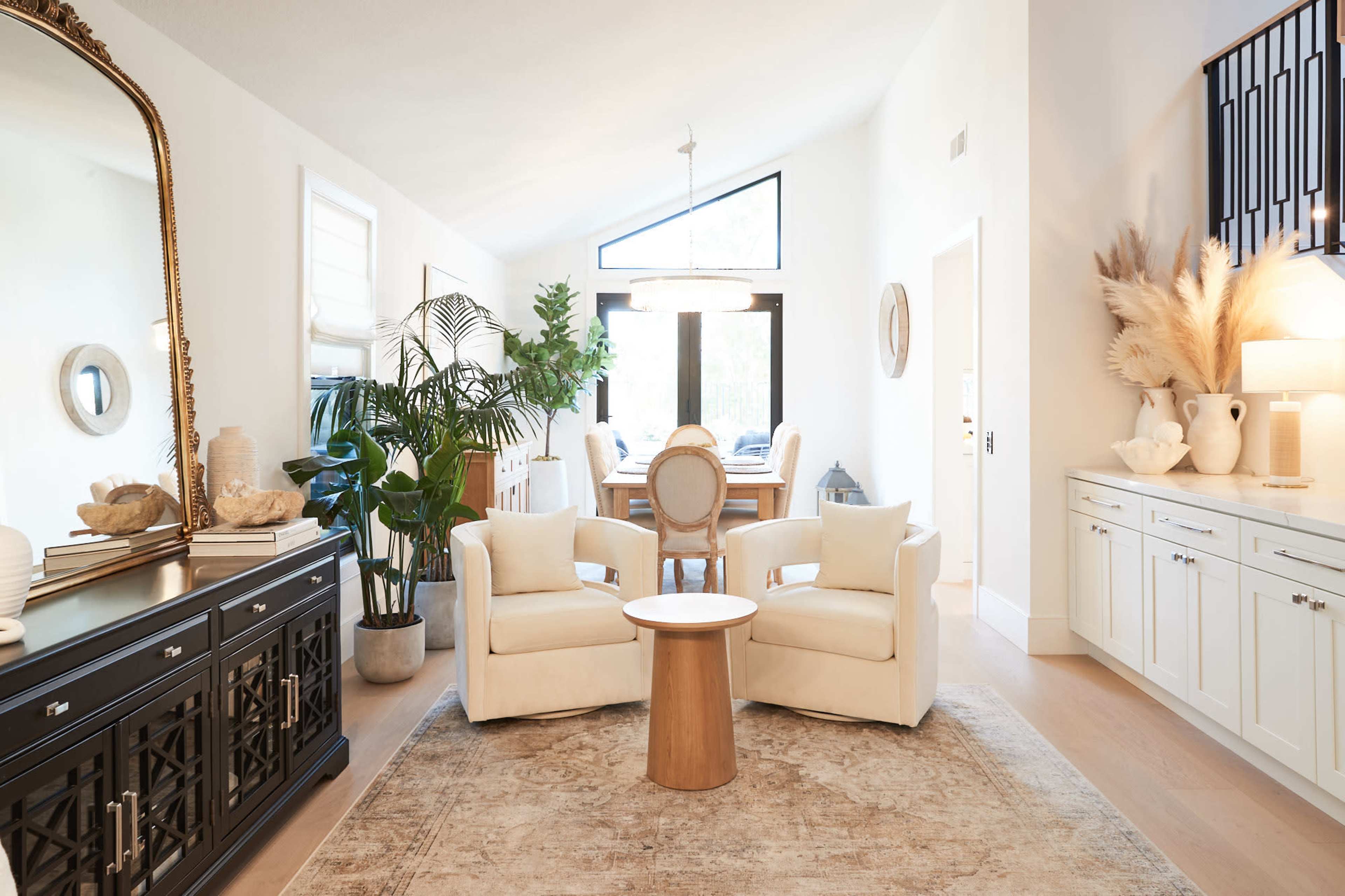 A bright living room featuring two white chairs, a small wooden table, a large wall mirror, and various decorative elements on a console table.