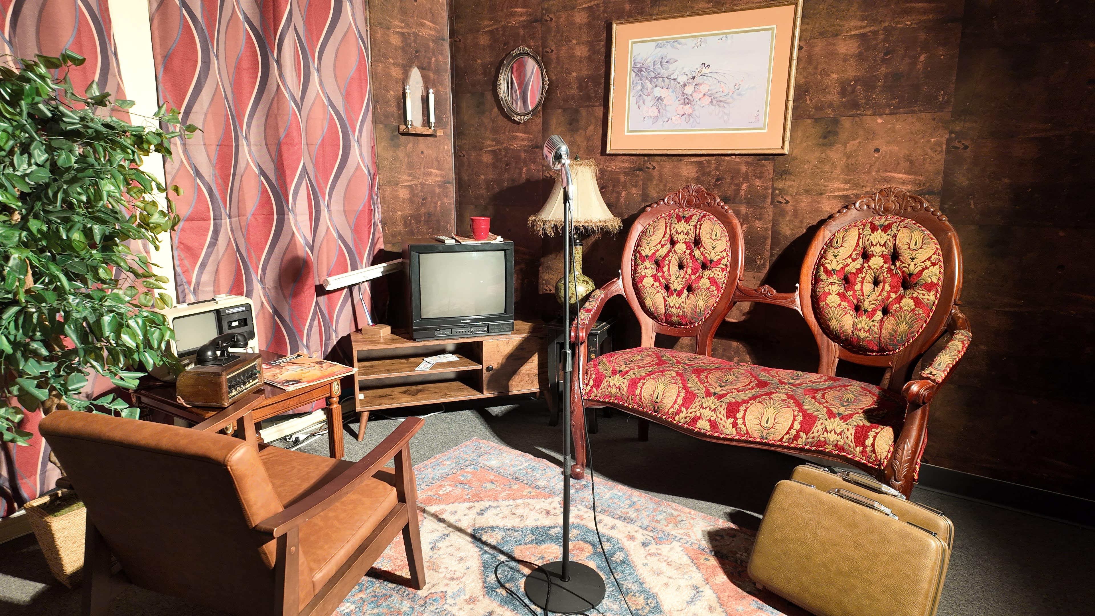The scene features a vintage-style living room with a patterned sofa, an old television, a microphone on a stand, and a suitcase placed on the floor.