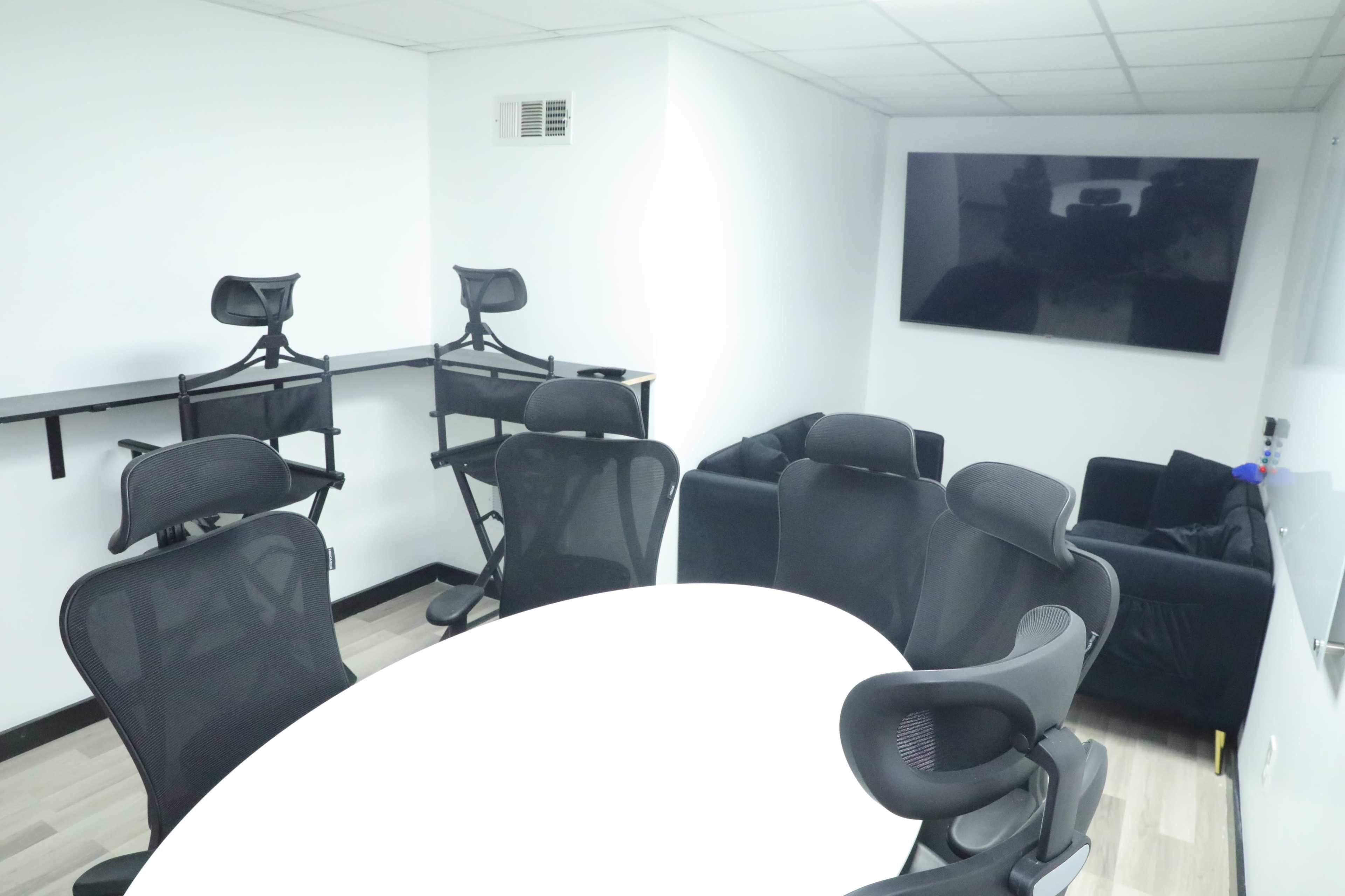 A modern meeting room with a white oval table, black chairs, and a wall-mounted flat-screen TV.