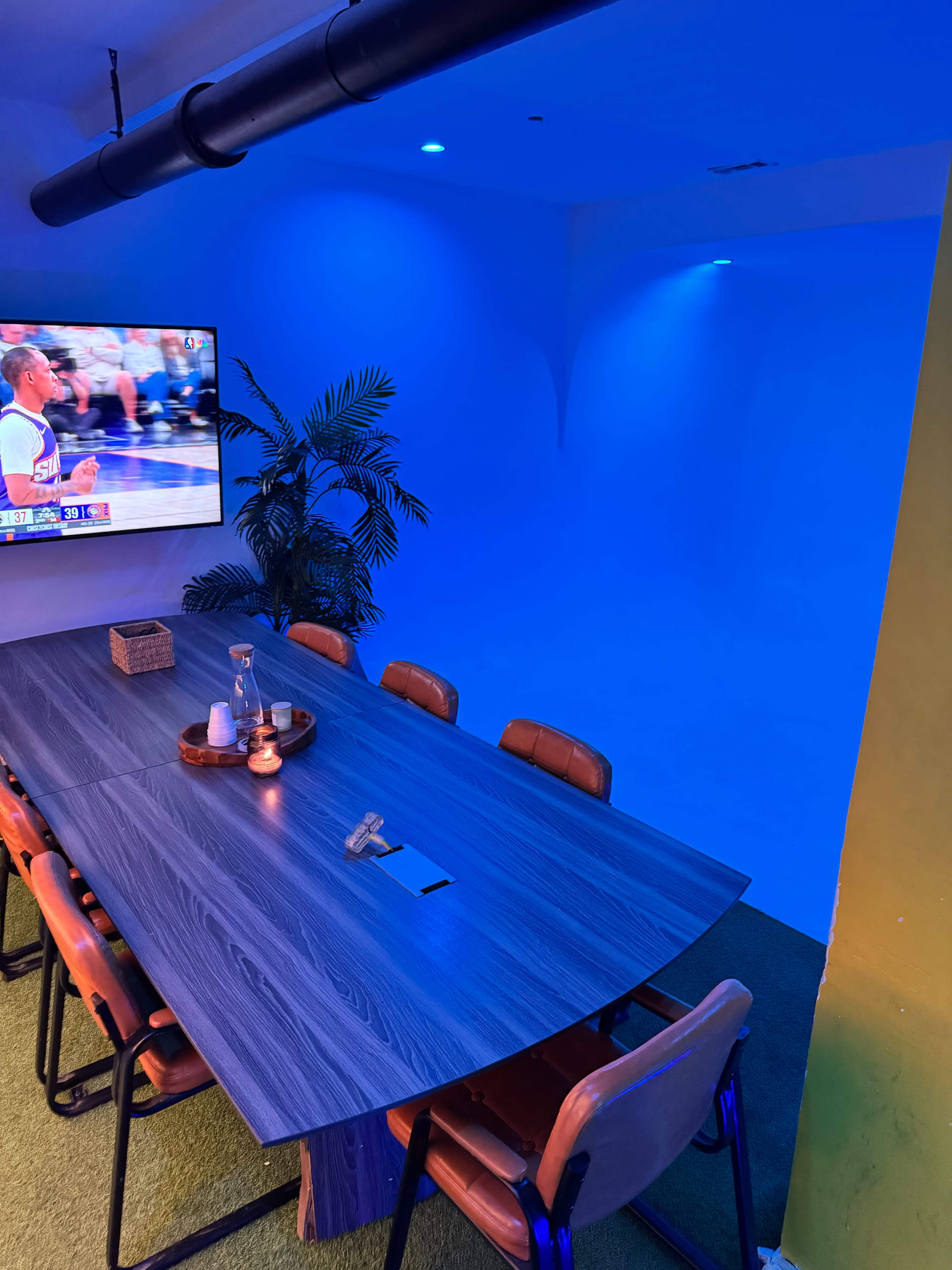 A large conference table with leather chairs is positioned in a room illuminated by blue lights, featuring a television displaying a basketball game.