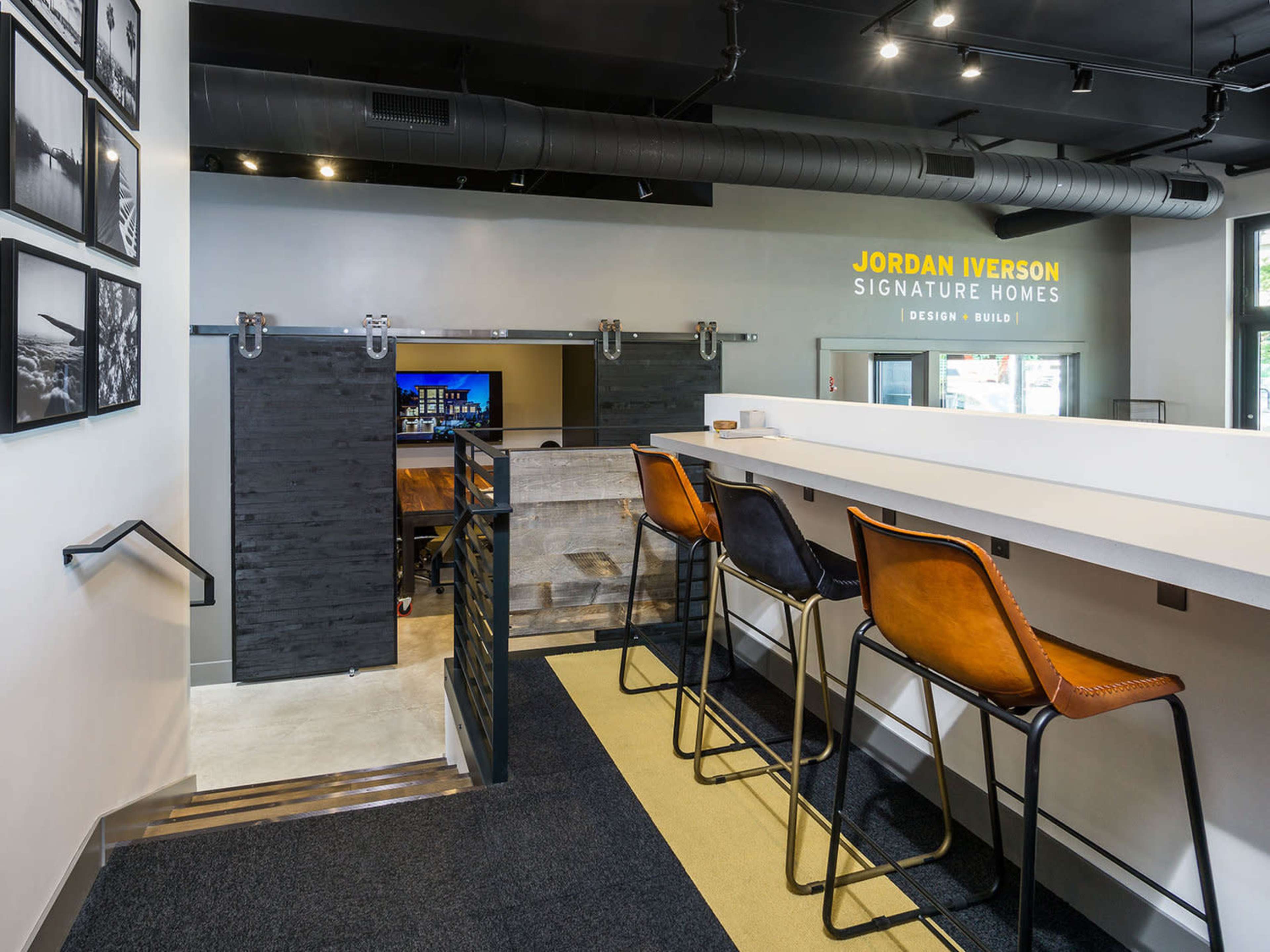 A modern interior space featuring a bar-style seating area with high stools, leading to a staircase and a large wall sign for "Jordan Iverson Signature Homes."