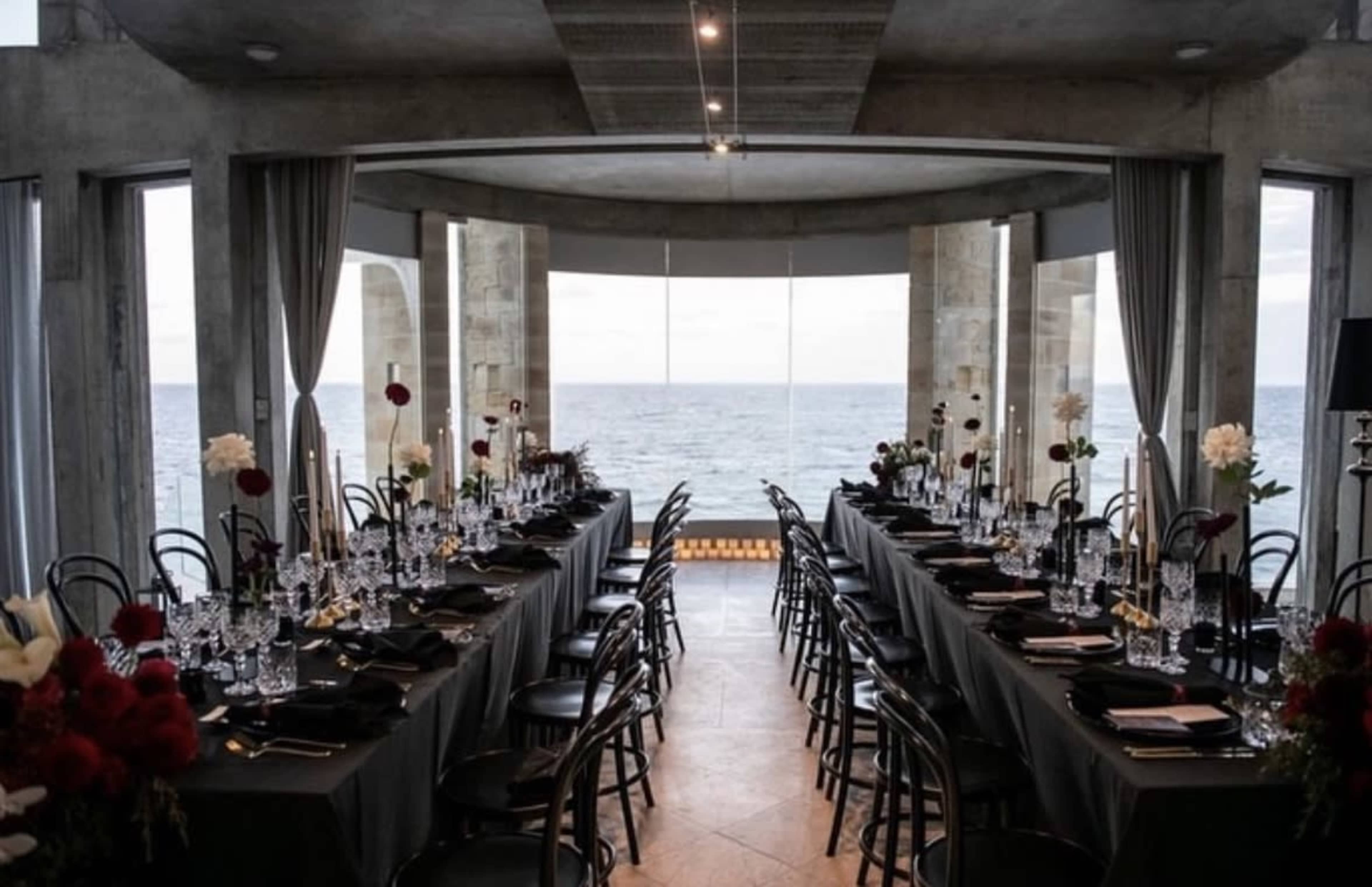 A long dining table is set for a formal meal with elegant tableware, facing large windows that overlook an ocean view.