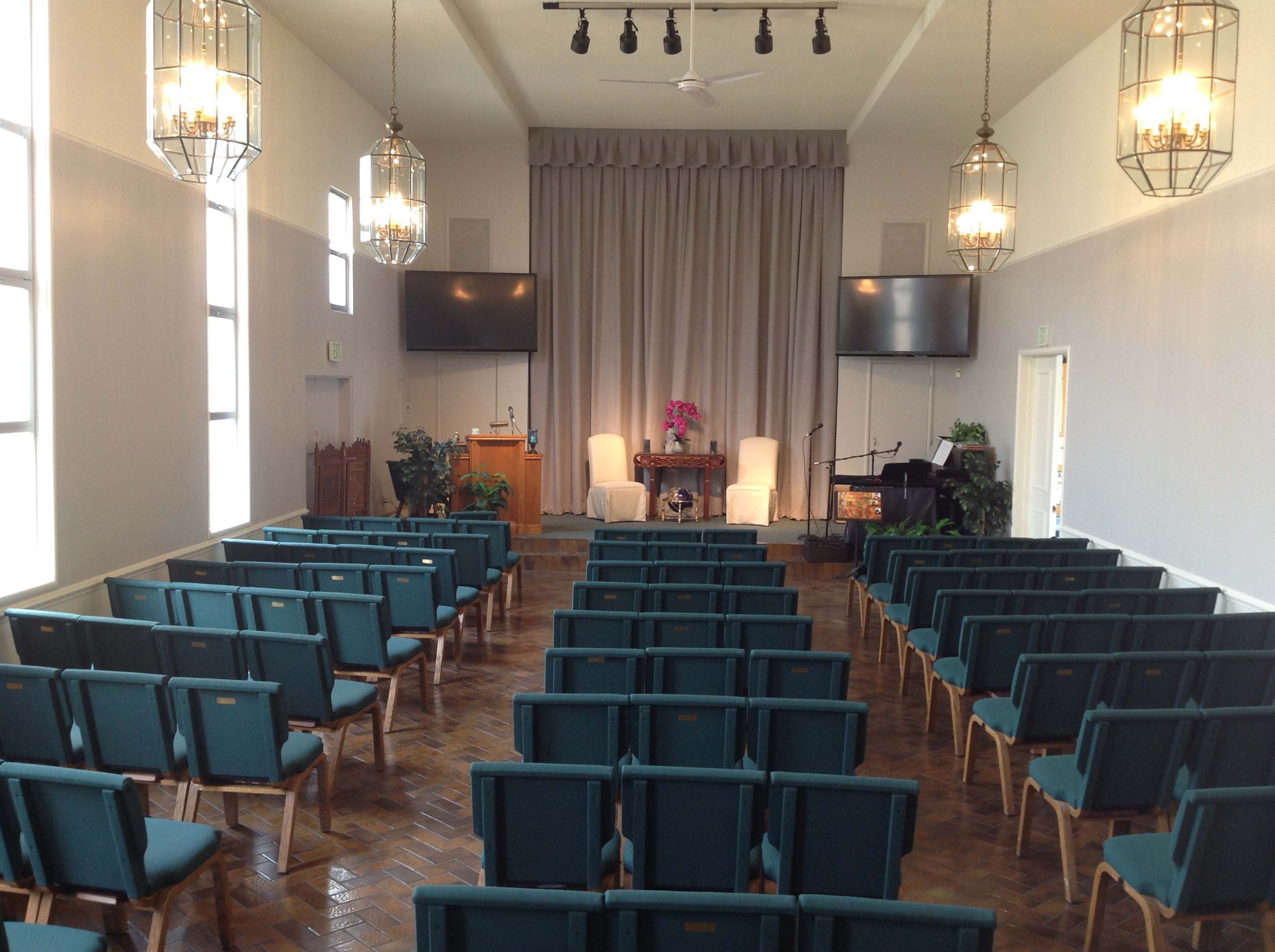 The interior of a venue features rows of teal chairs facing a small stage with two white armchairs, a microphone, and a piano, all framed by large windows and pendant lights.
