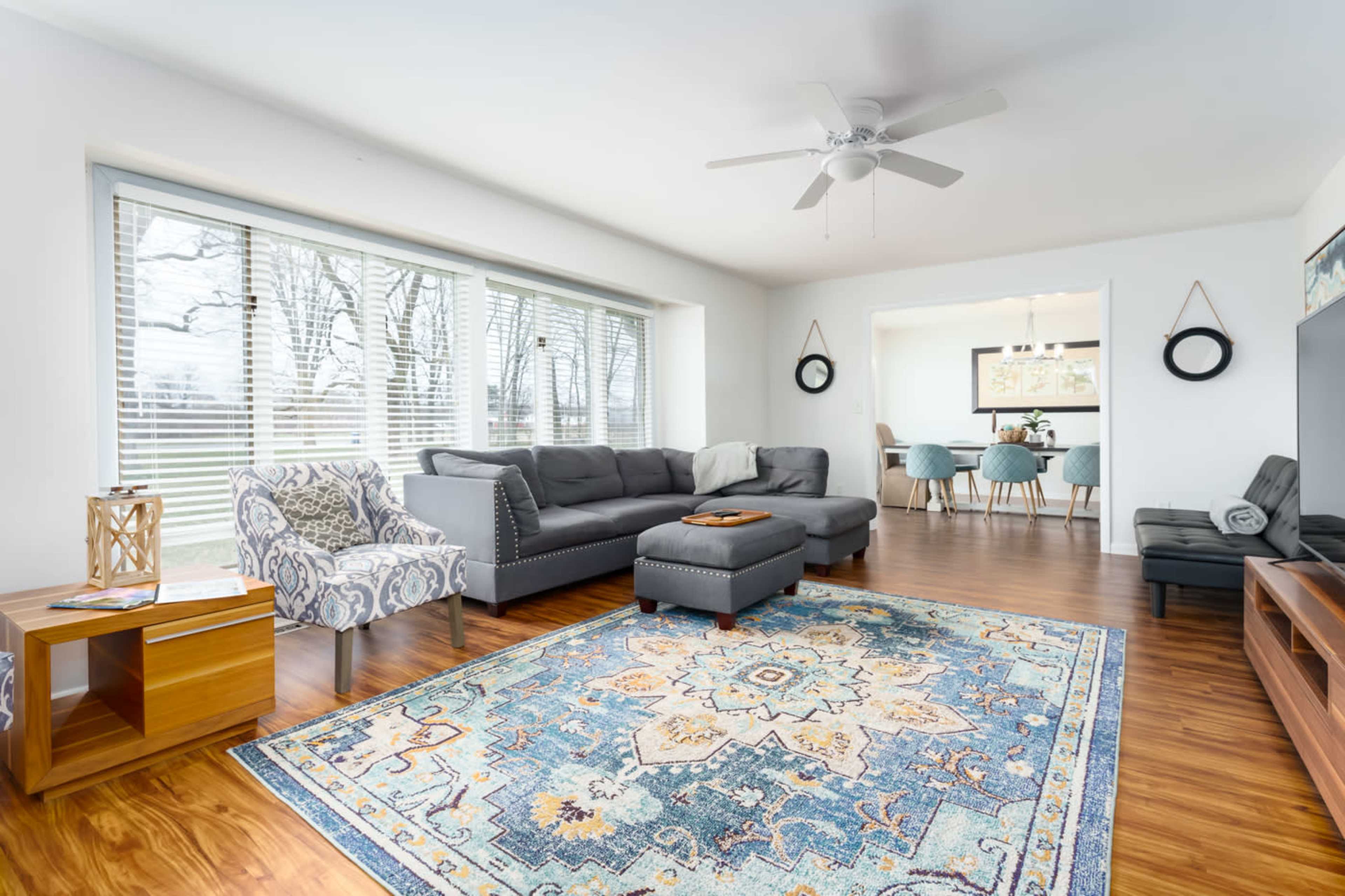 The room features a modern living area with a large sectional couch, a patterned area rug, and a view of trees through expansive windows.