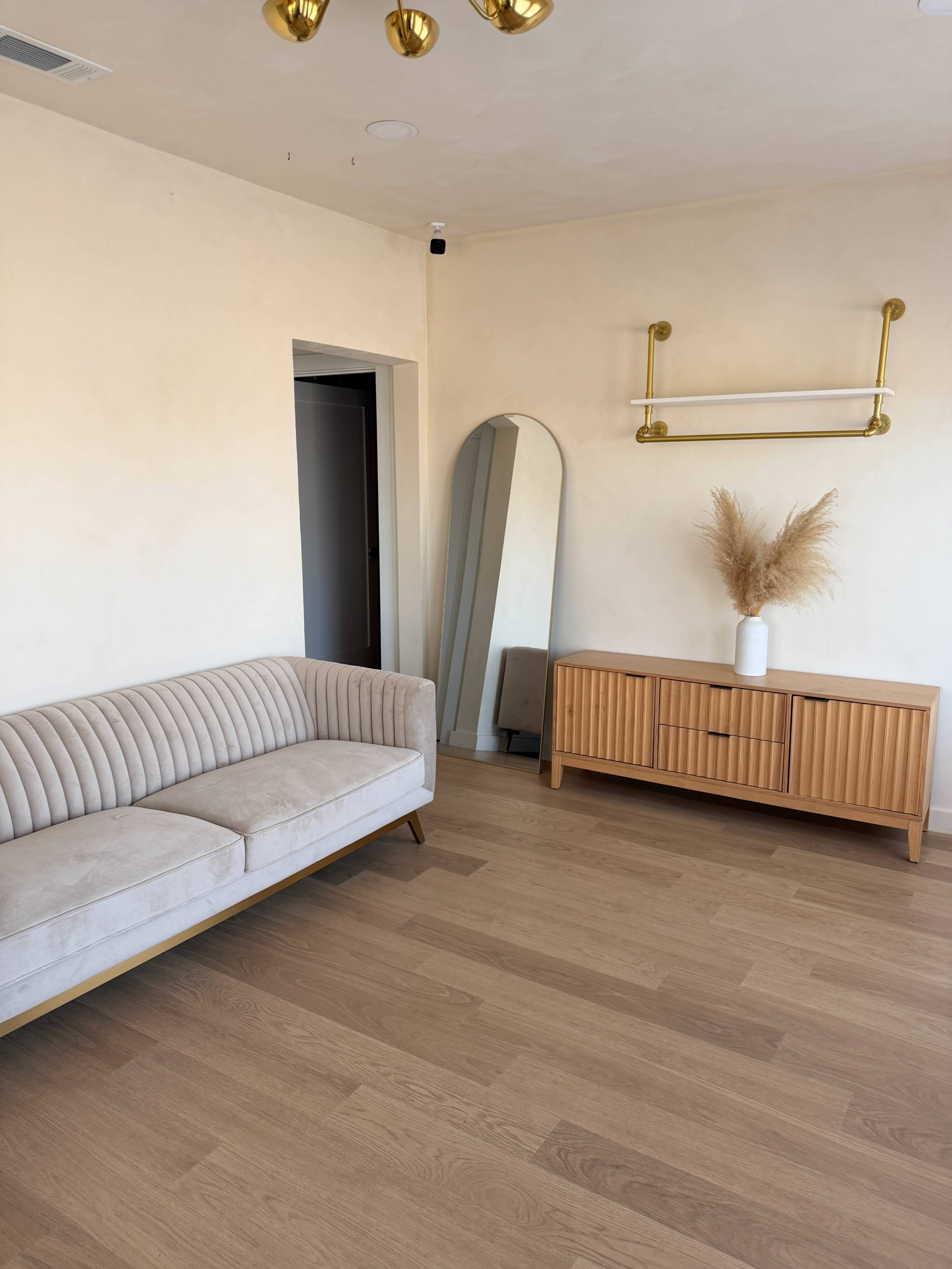 The image shows a simple room with a beige sofa, a wooden cabinet, and a large mirror against a light-colored wall.