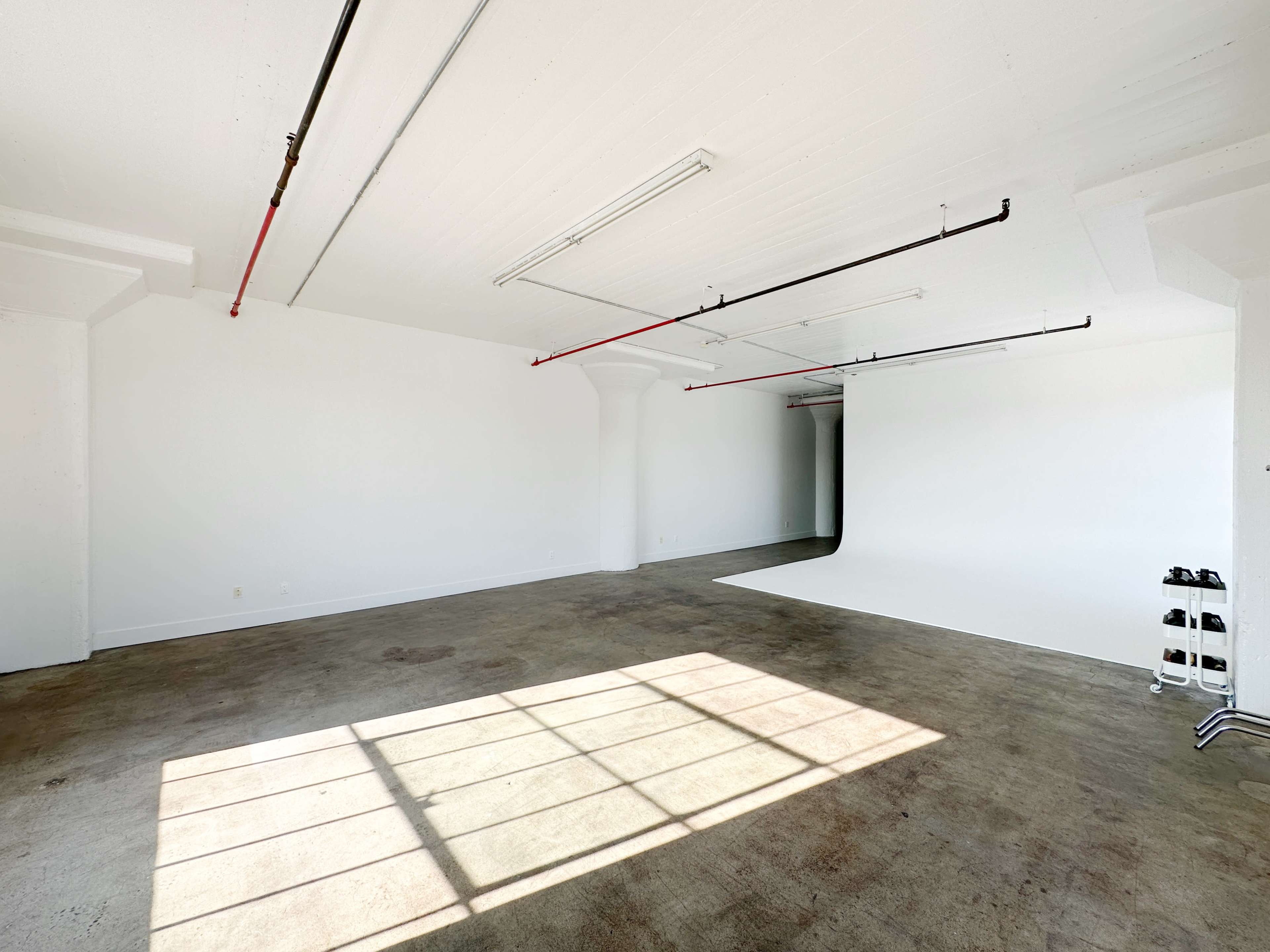 A spacious, empty room with white walls, a concrete floor, and natural light entering through a large window.