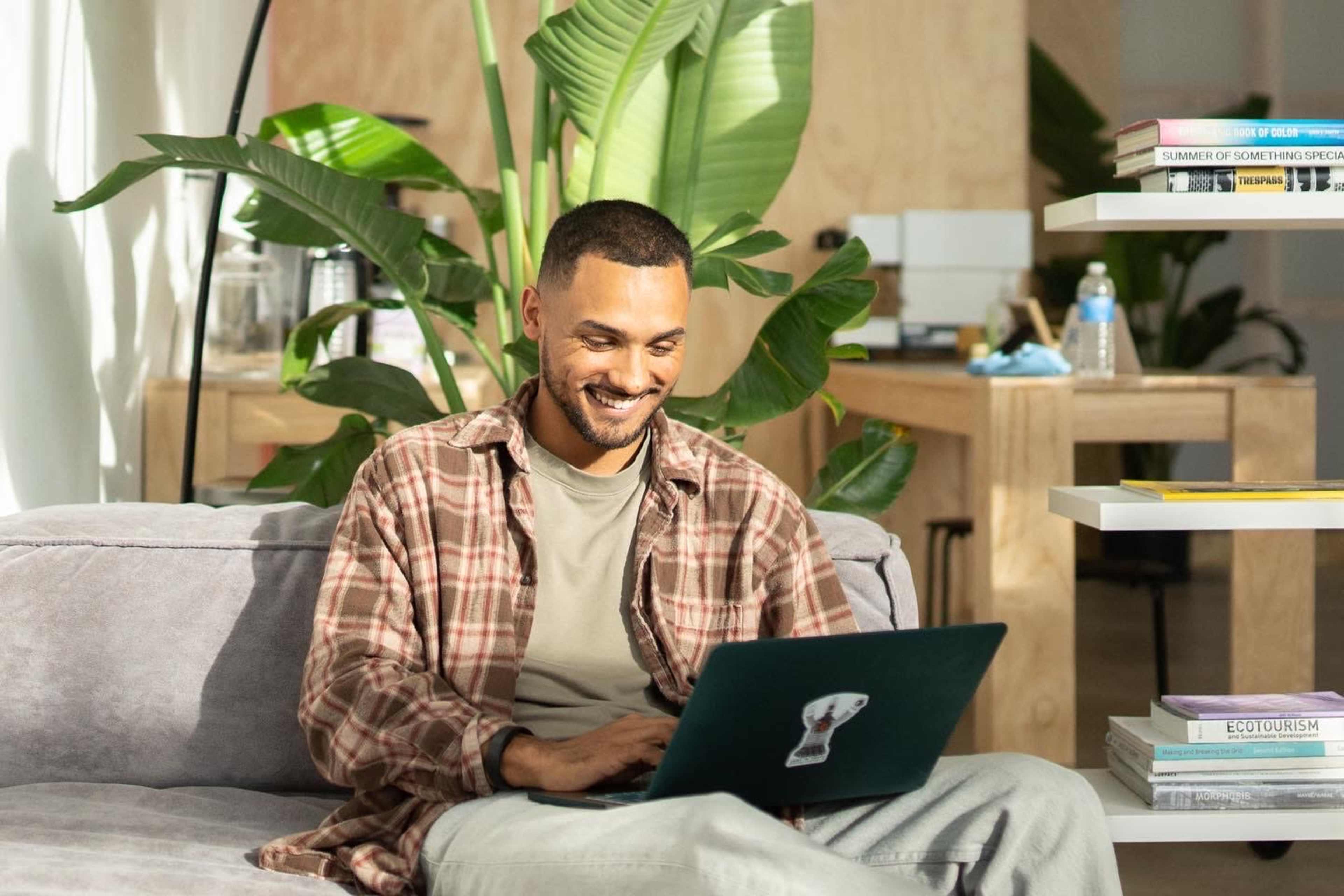 A man sits on a gray couch, working on a laptop in a bright room filled with plants and a bookshelf.