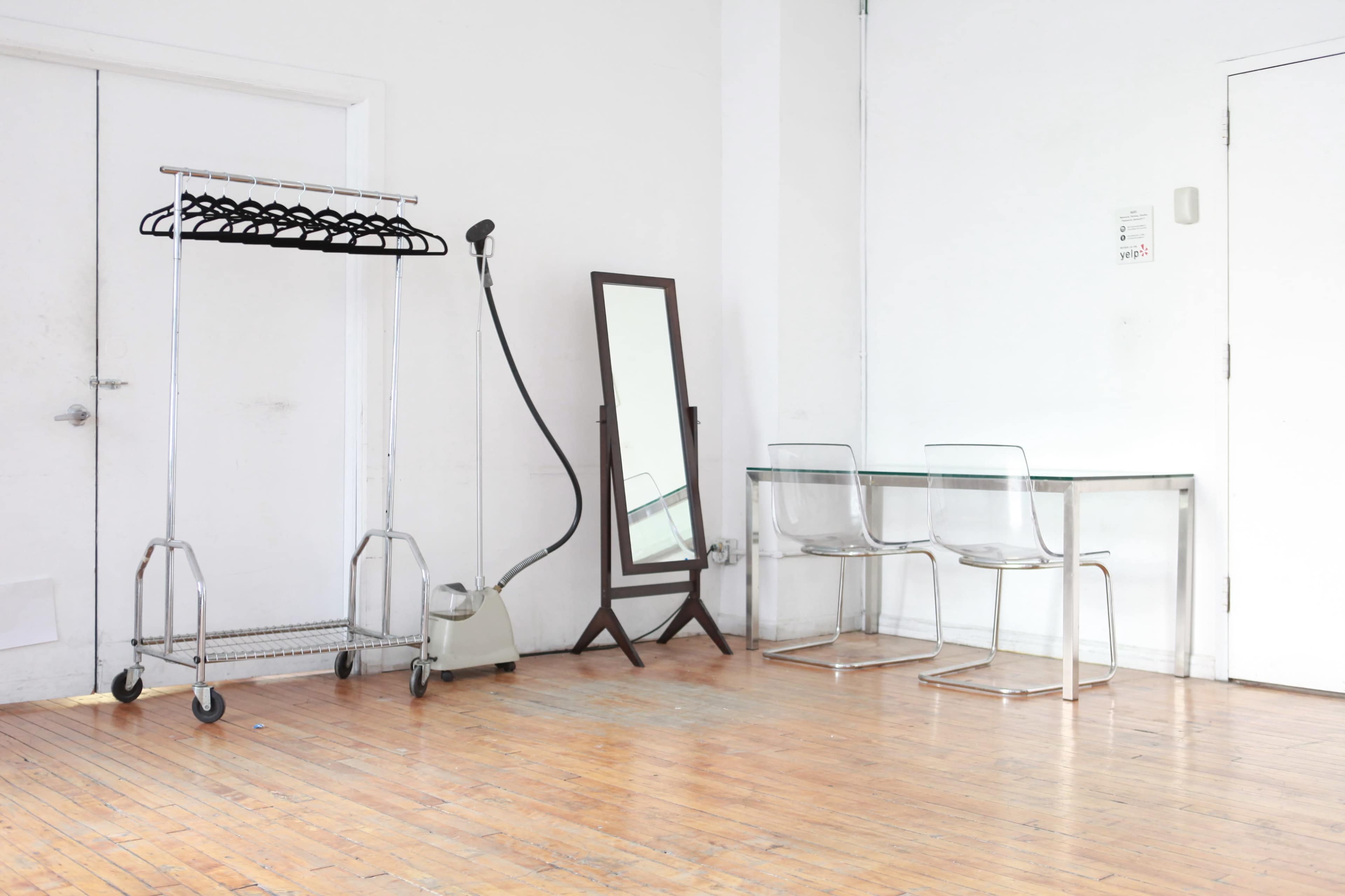 The image depicts a minimalist interior space featuring a clothing rack, a steam cleaner, a full-length mirror, and two clear chairs arranged around a glass table on a wooden floor.