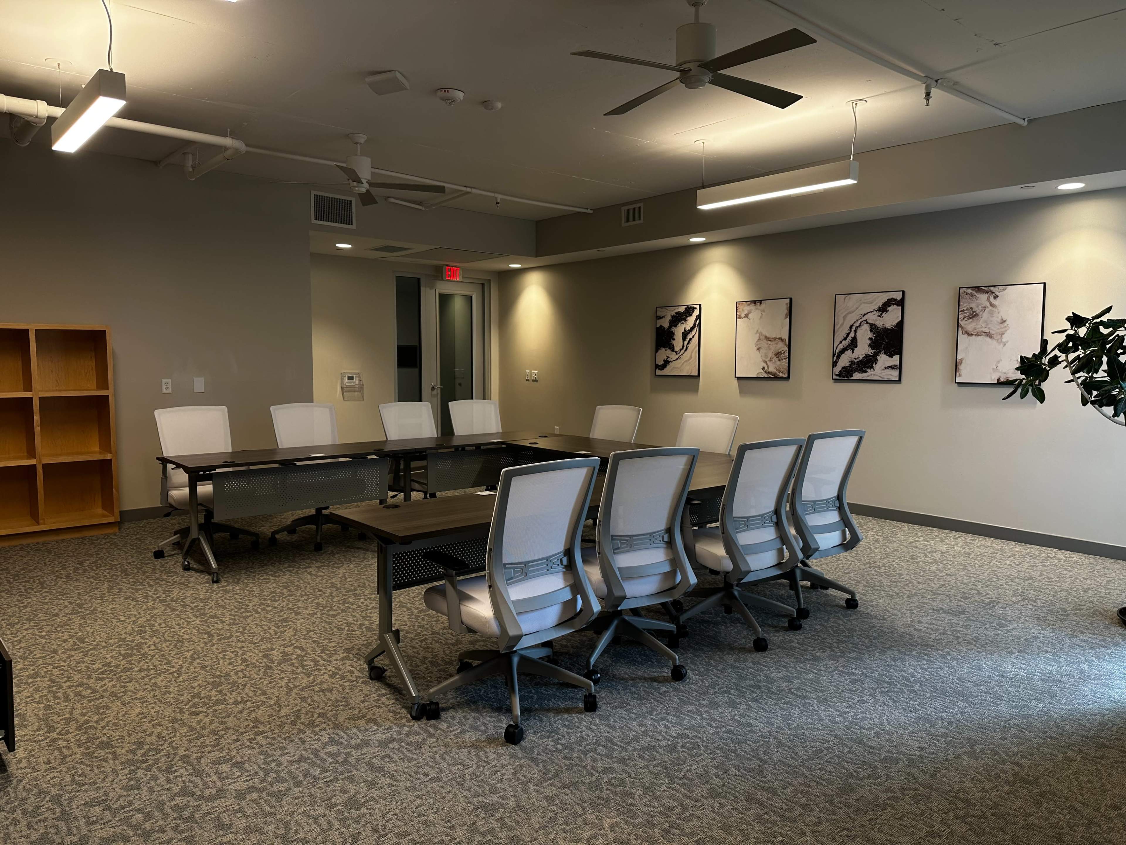 A modern conference room features a long table surrounded by several ergonomic chairs, with artwork on the walls and a bookshelf in one corner.