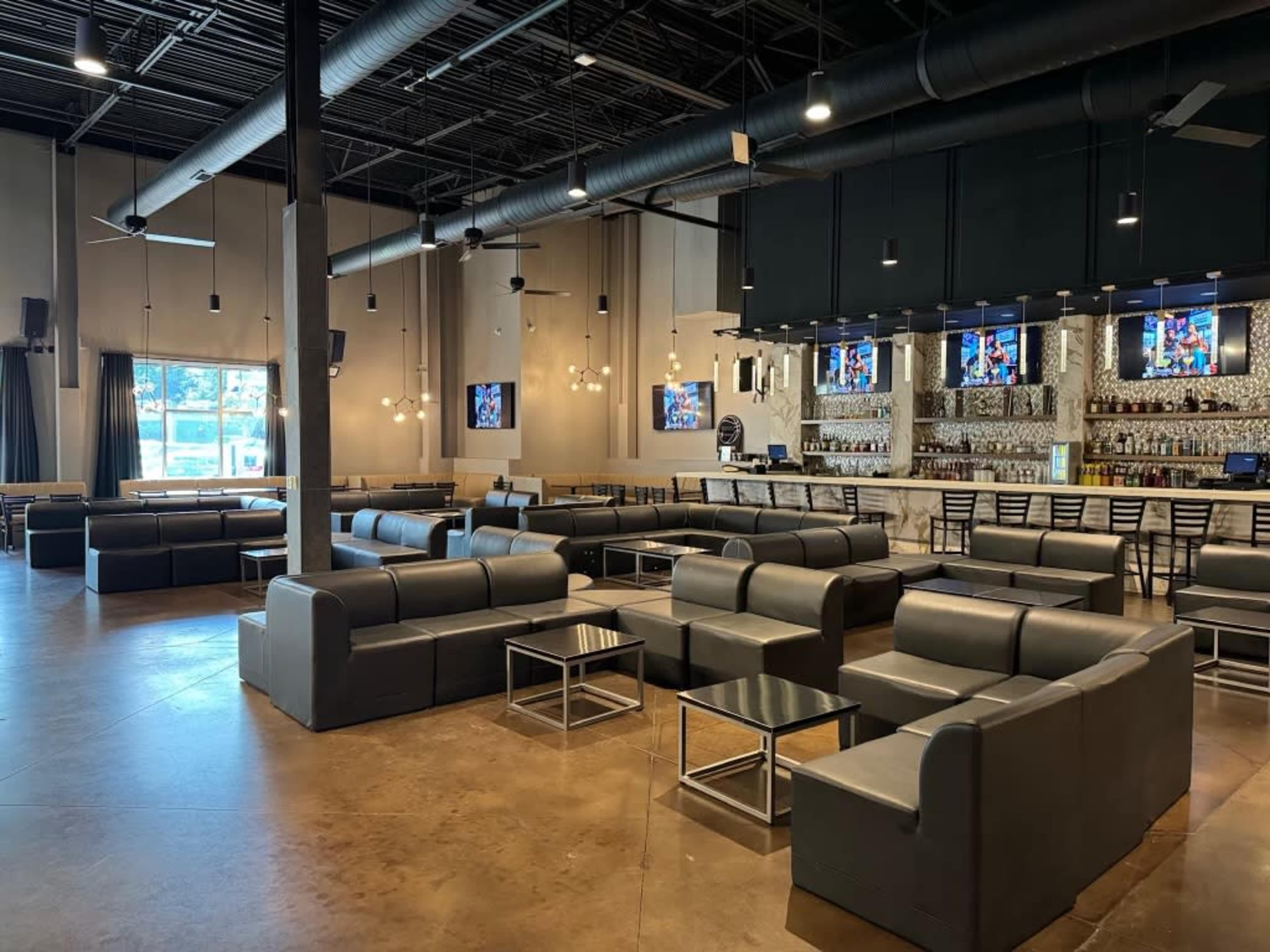 A spacious, modern lounge area with gray sofas and a bar counter featuring multiple flat-screen TVs in a well-lit interior.