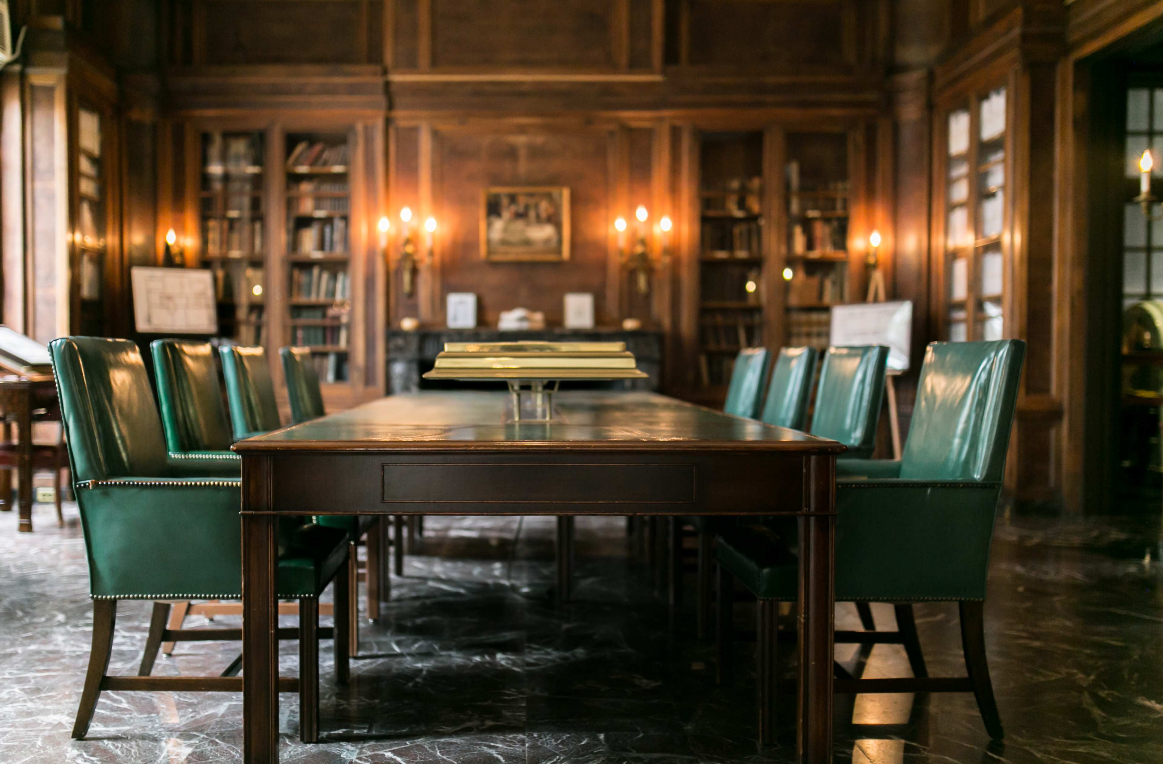 A long wooden table surrounded by green leather chairs is set in a dimly lit library filled with bookshelves and ornate lighting.