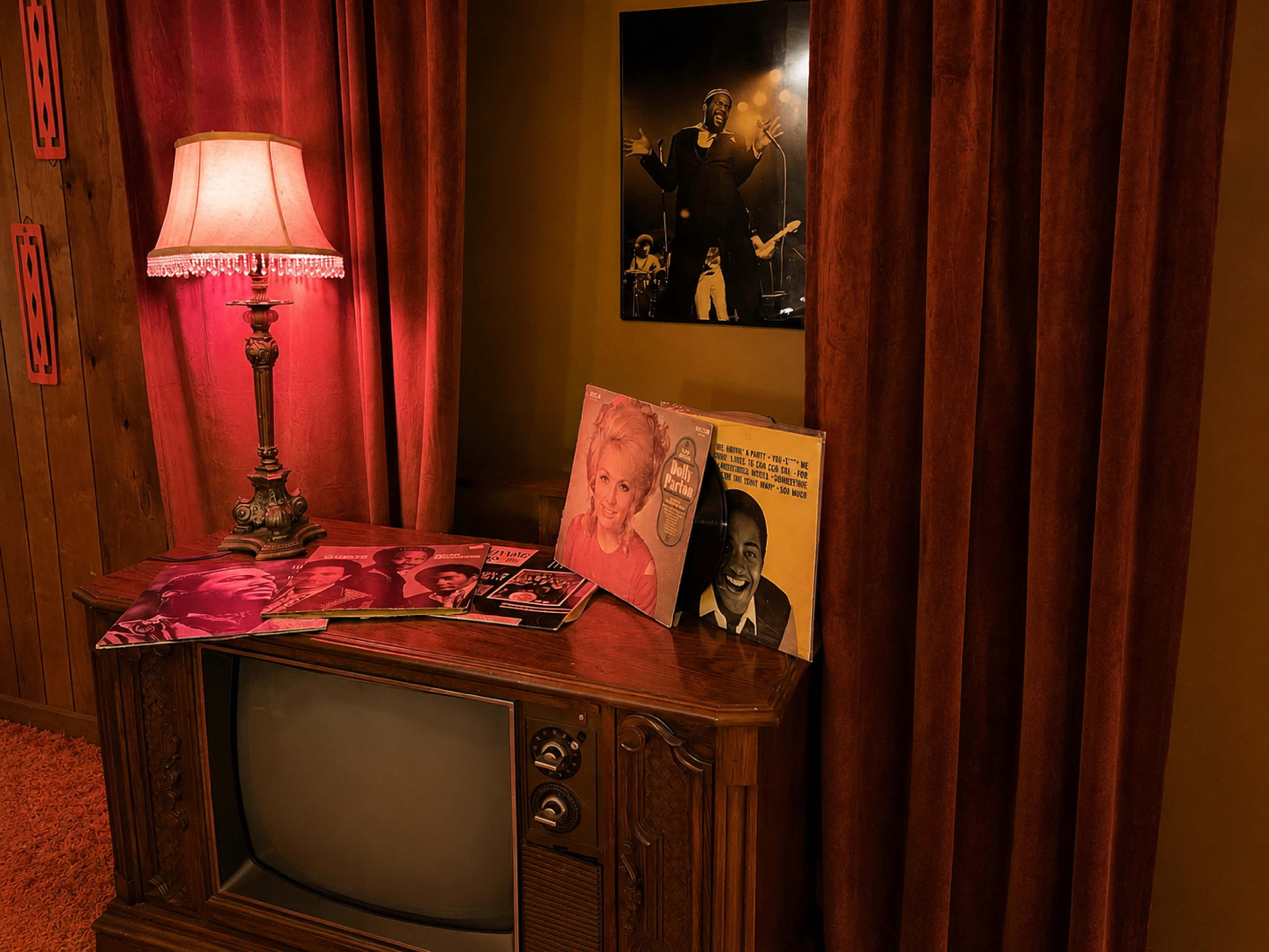 A vintage television set sits on a wooden table adorned with vinyl records, beside a lamp casting a warm glow in a room with red curtains and wood-paneled walls.