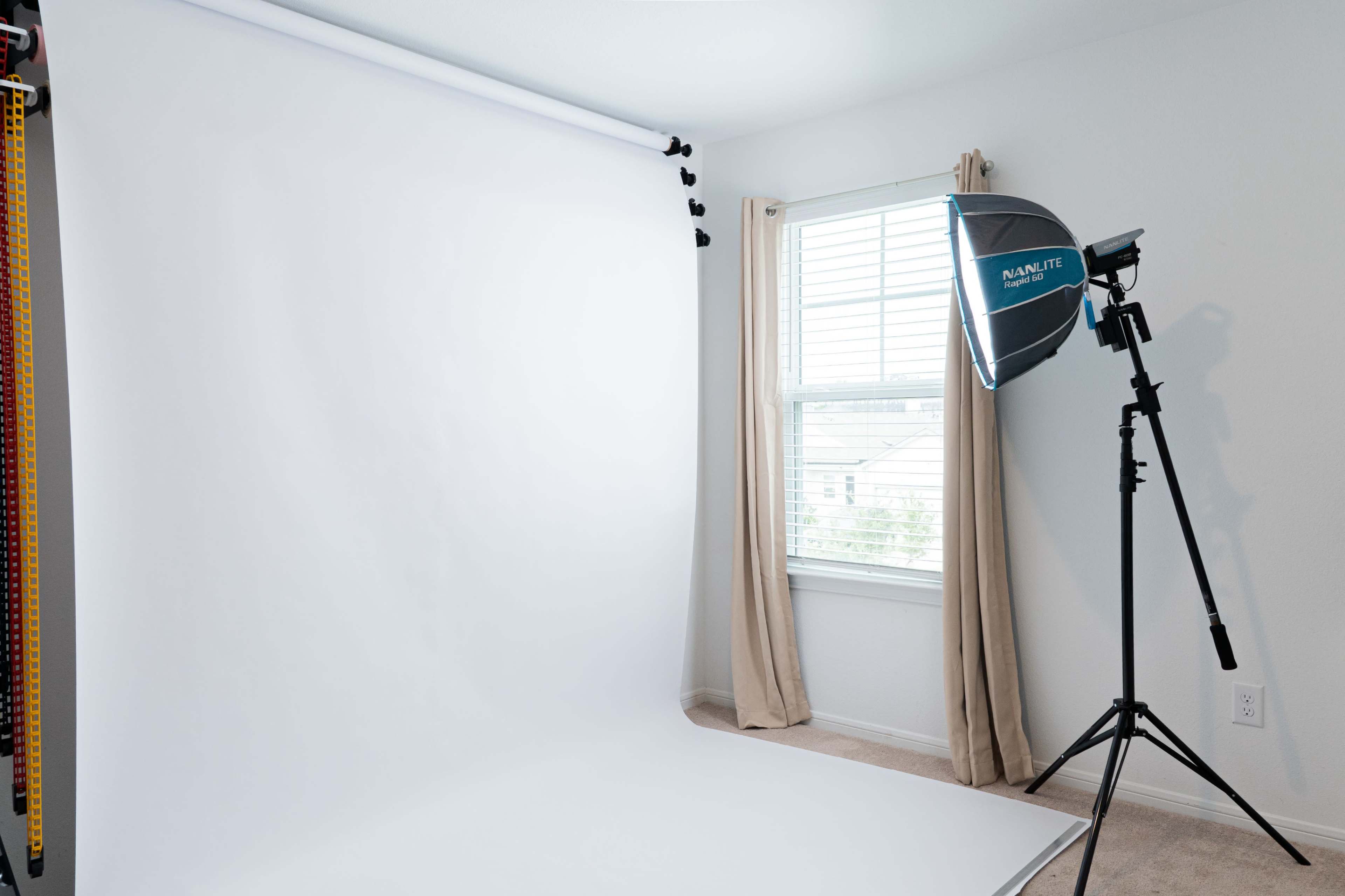 A photography studio setup features a seamless white backdrop, soft natural light from a window, and a mounted light source.