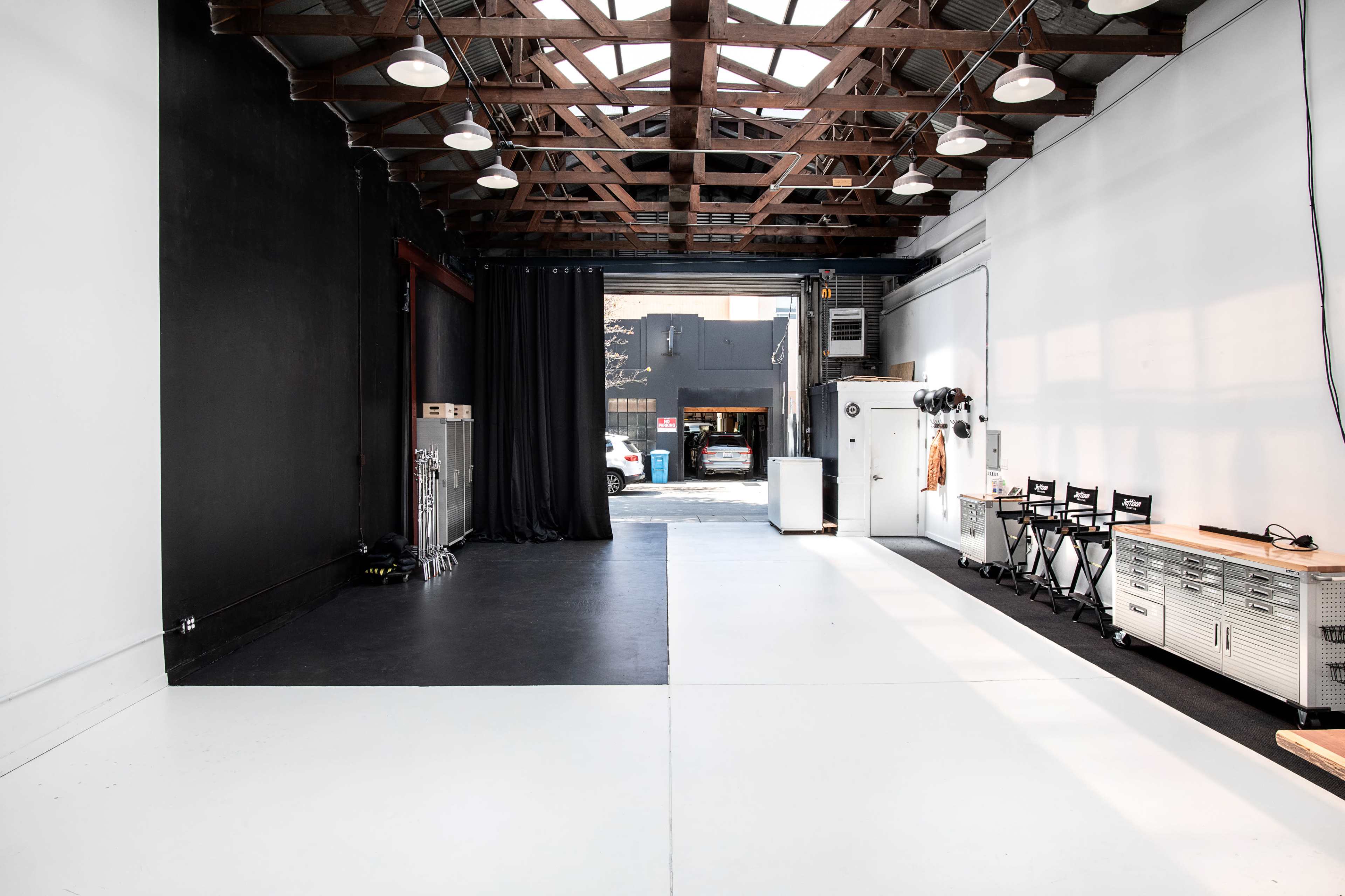 A large, open space featuring a black wall on one side and a white wall on the other, with a ceiling supported by wooden beams and a small parking area visible through a doorway.