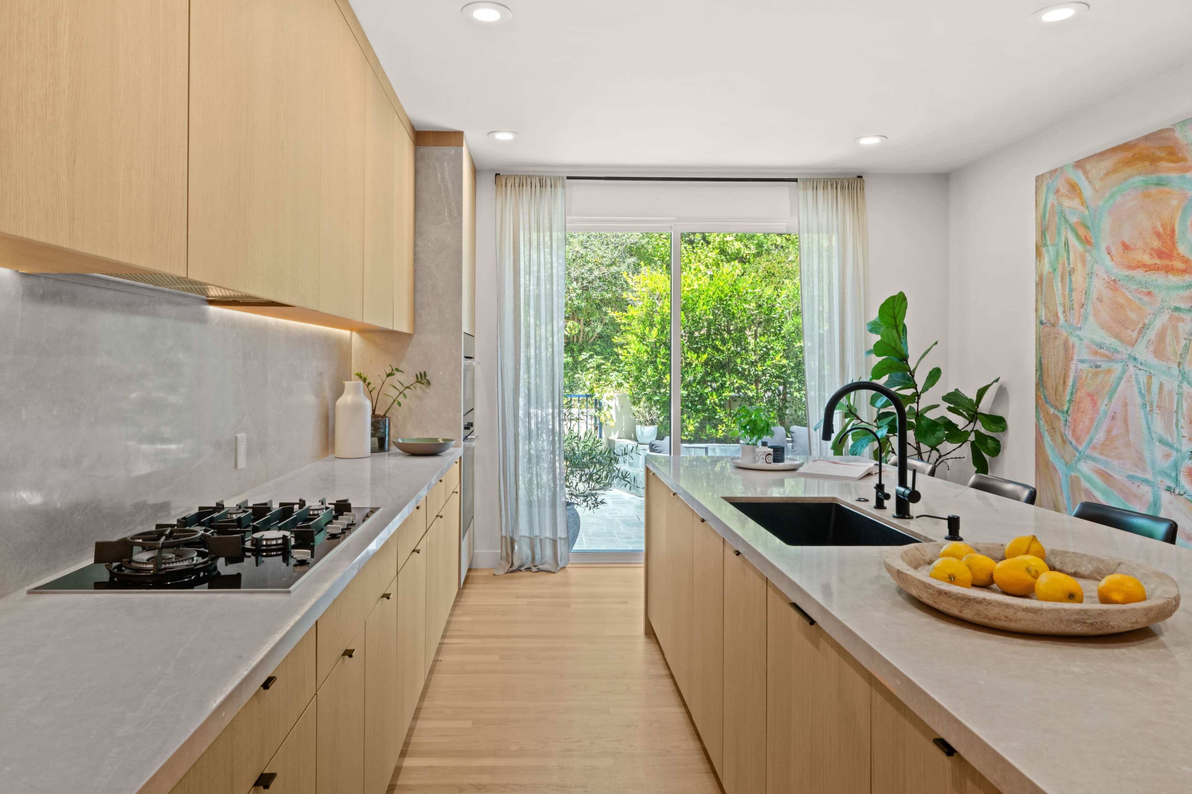 The image shows a modern kitchen with light wooden cabinetry, a large island with a sink, and a sliding glass door that opens to a garden area.