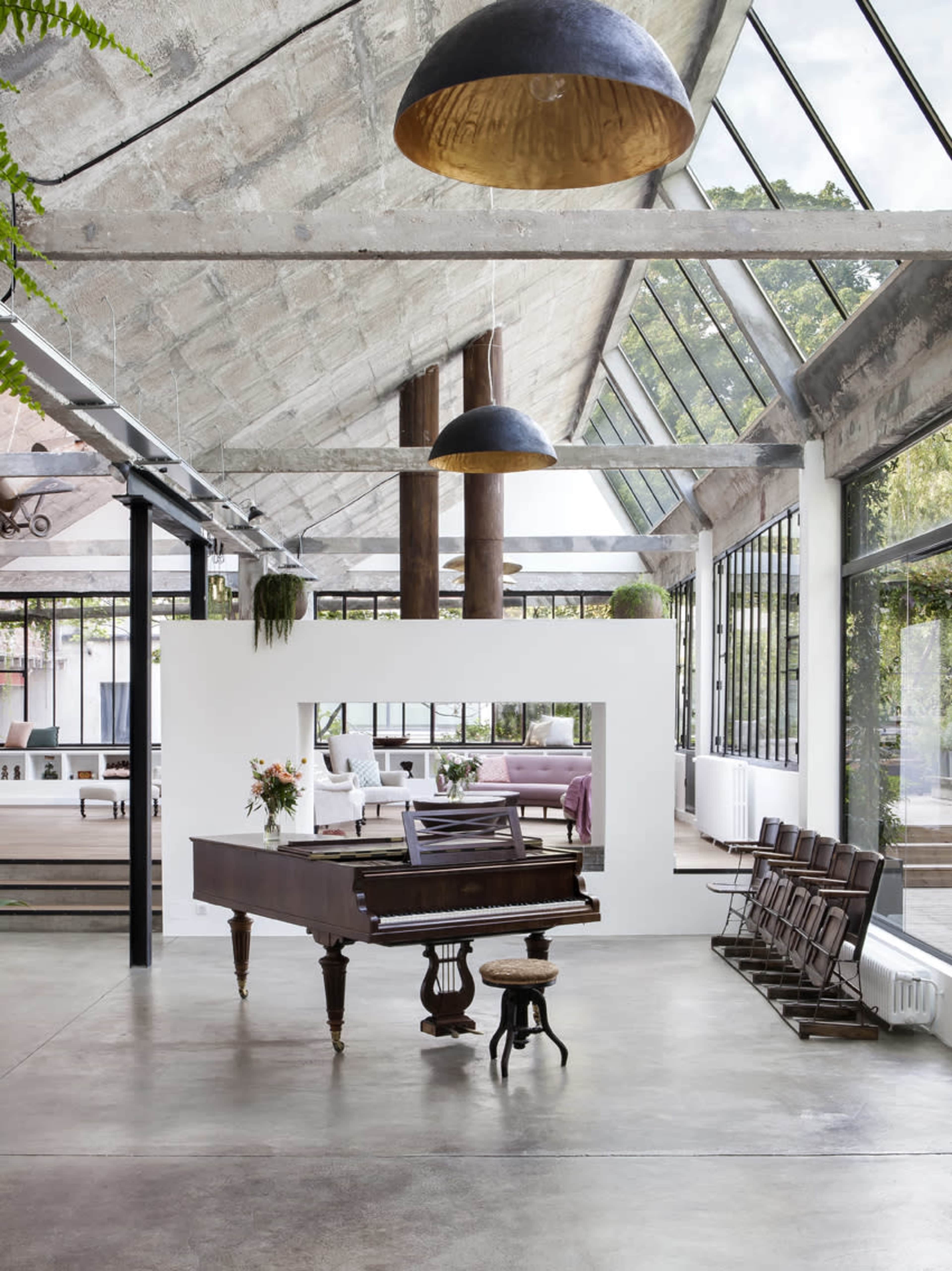 A grand piano is positioned in the center of a spacious room with high ceilings, large windows, and industrial decor elements.