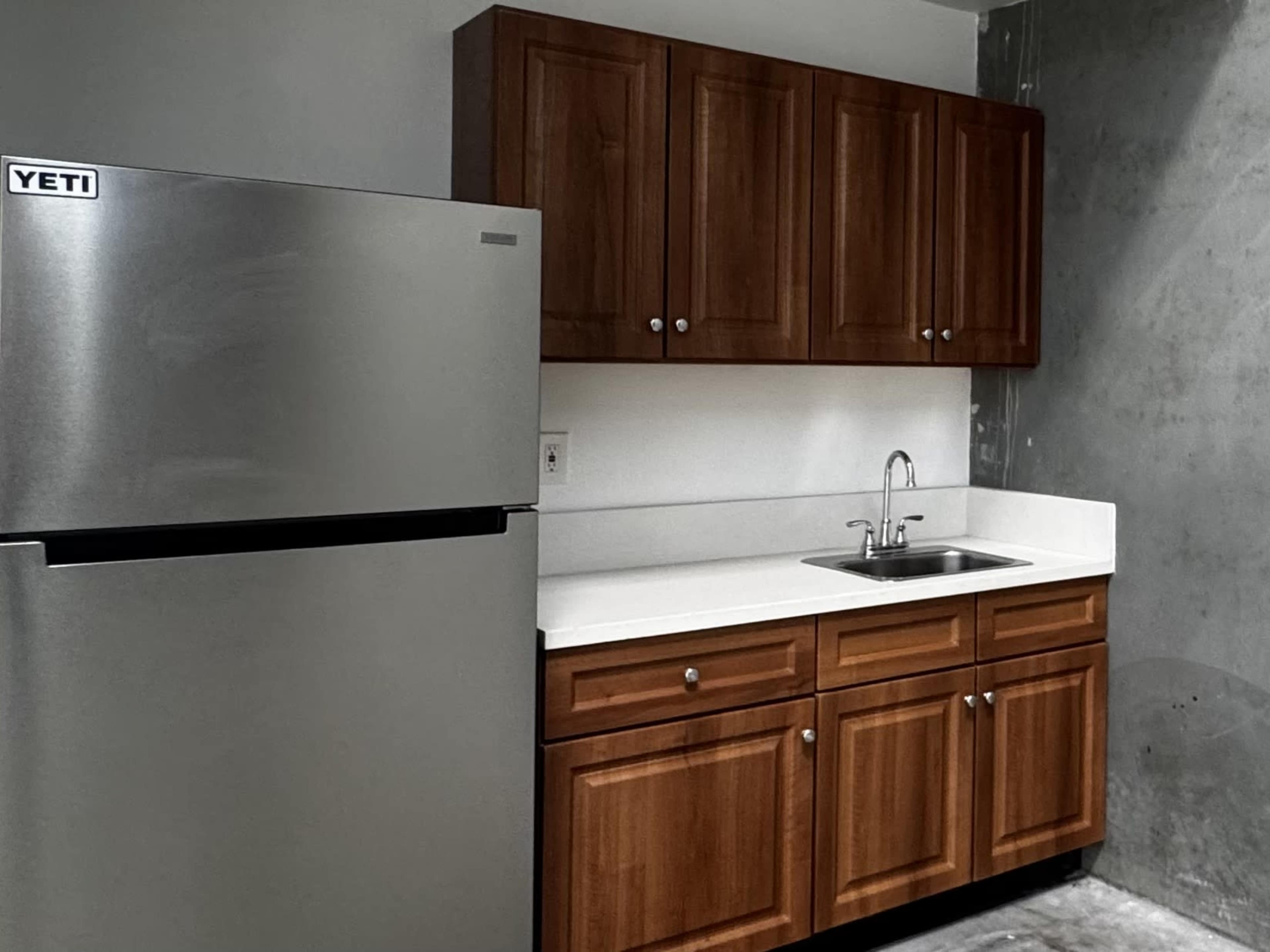 The image shows a kitchen space featuring a stainless steel refrigerator, wooden cabinets, and a small sink with a countertop.
