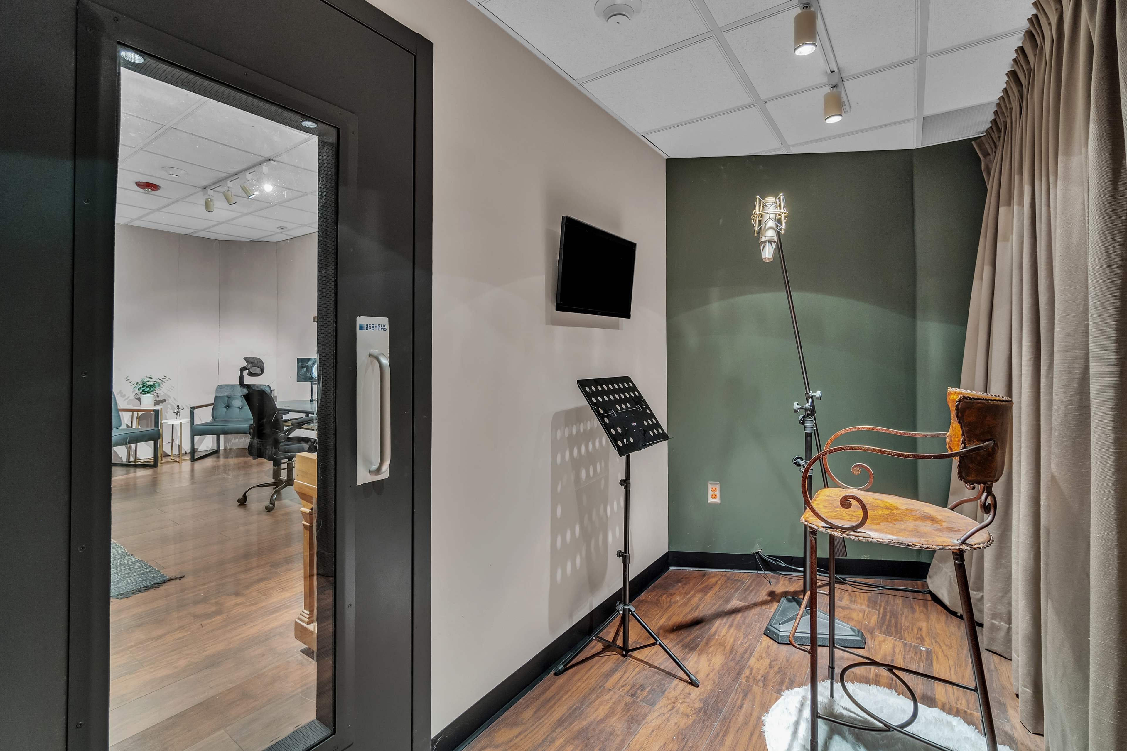 The image shows a sound booth with a microphone stand and an antique chair, visible through a glass door, leading to an office space in the background.