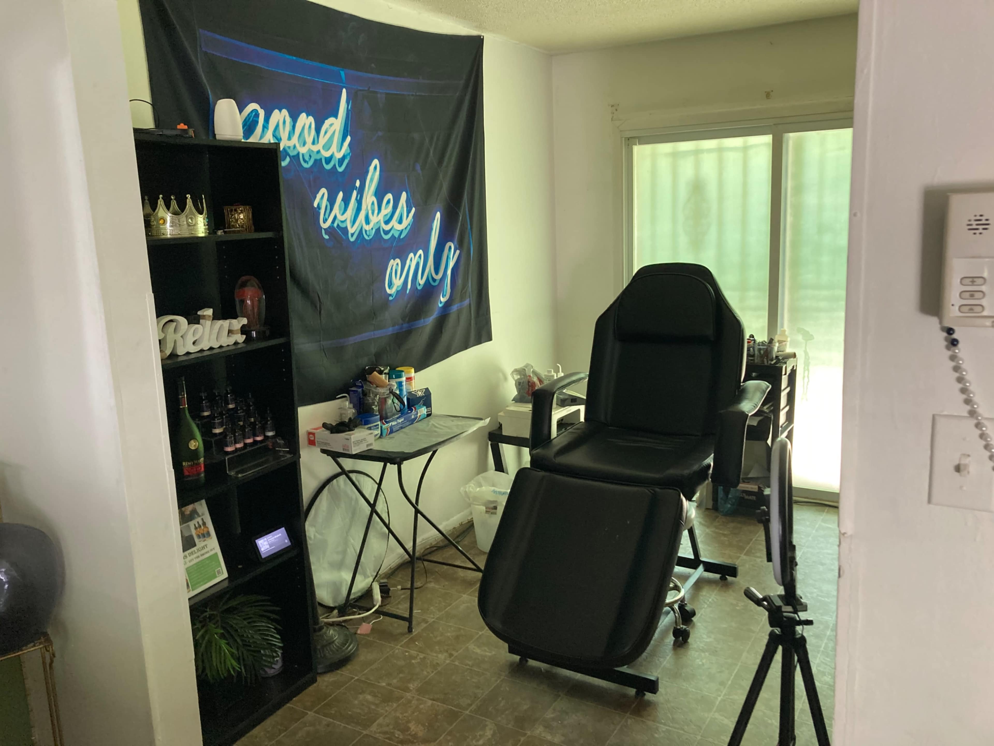 A small room with a tattoo chair, a table covered with supplies, a shelf of drinks, and a wall hanging reading "good vibes only."