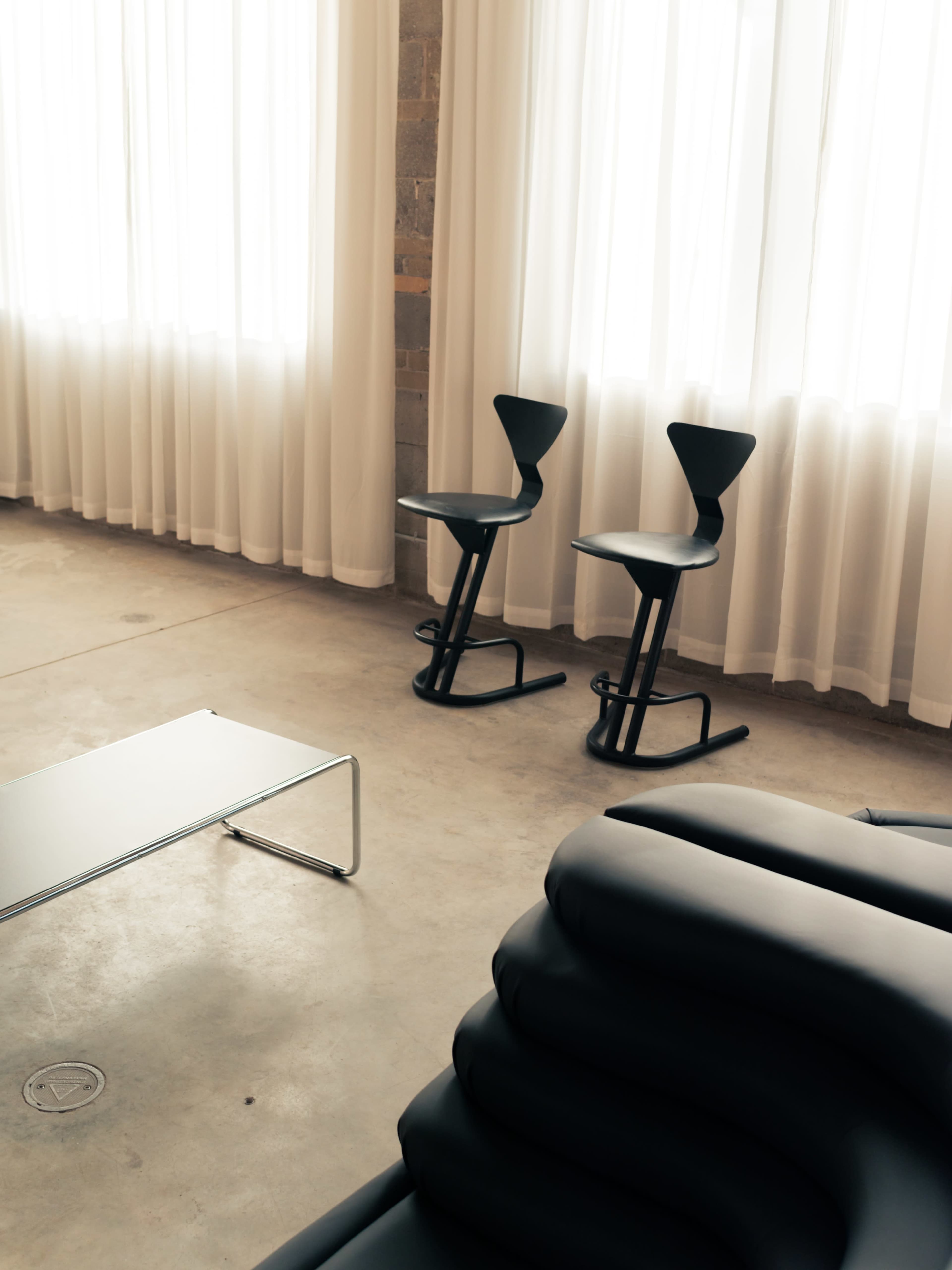 A minimalist interior features two black chairs and a low coffee table against a backdrop of sheer white curtains.
