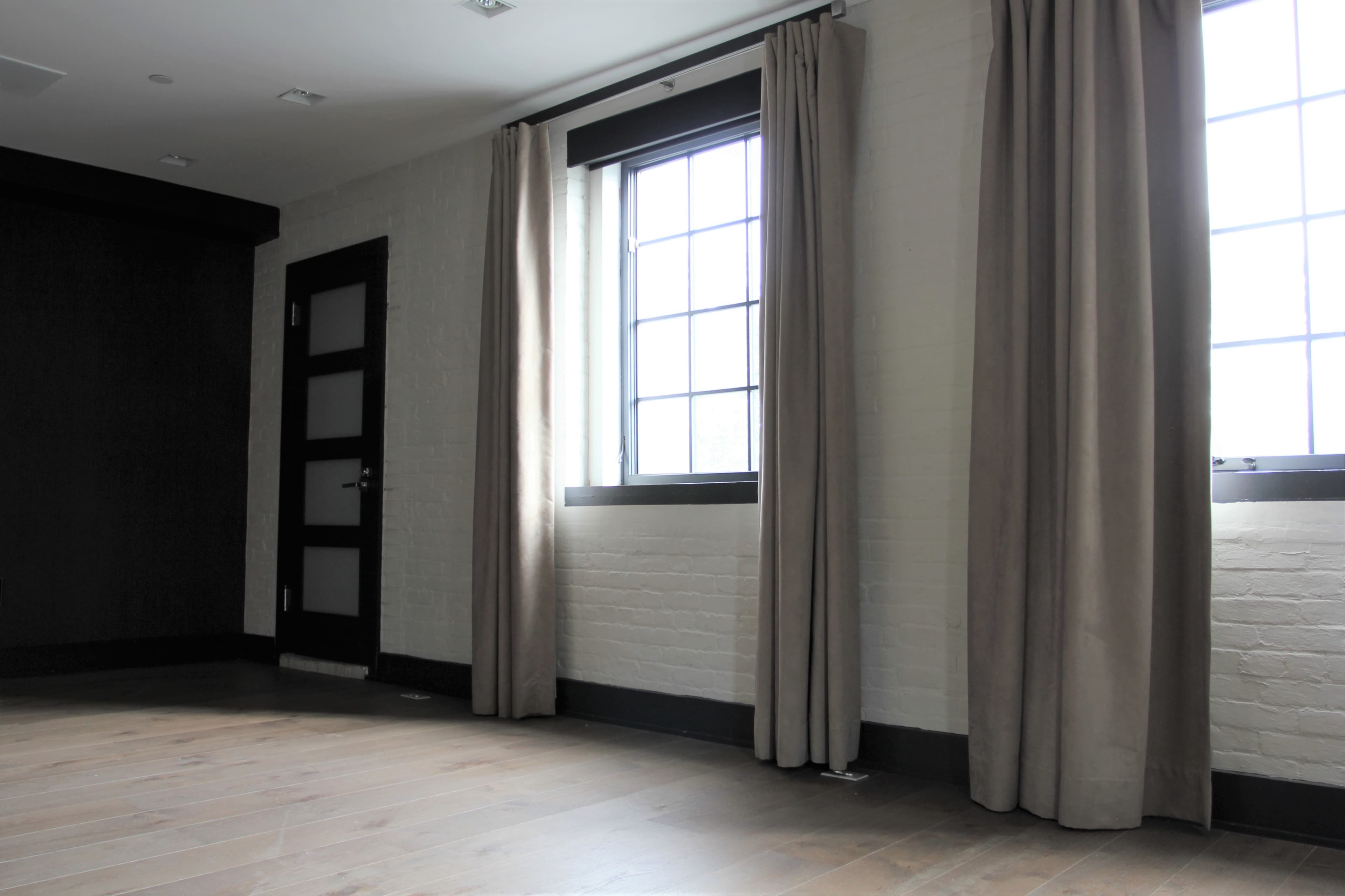 A spacious room with light-colored walls, wooden flooring, and large windows adorned with long, beige curtains.