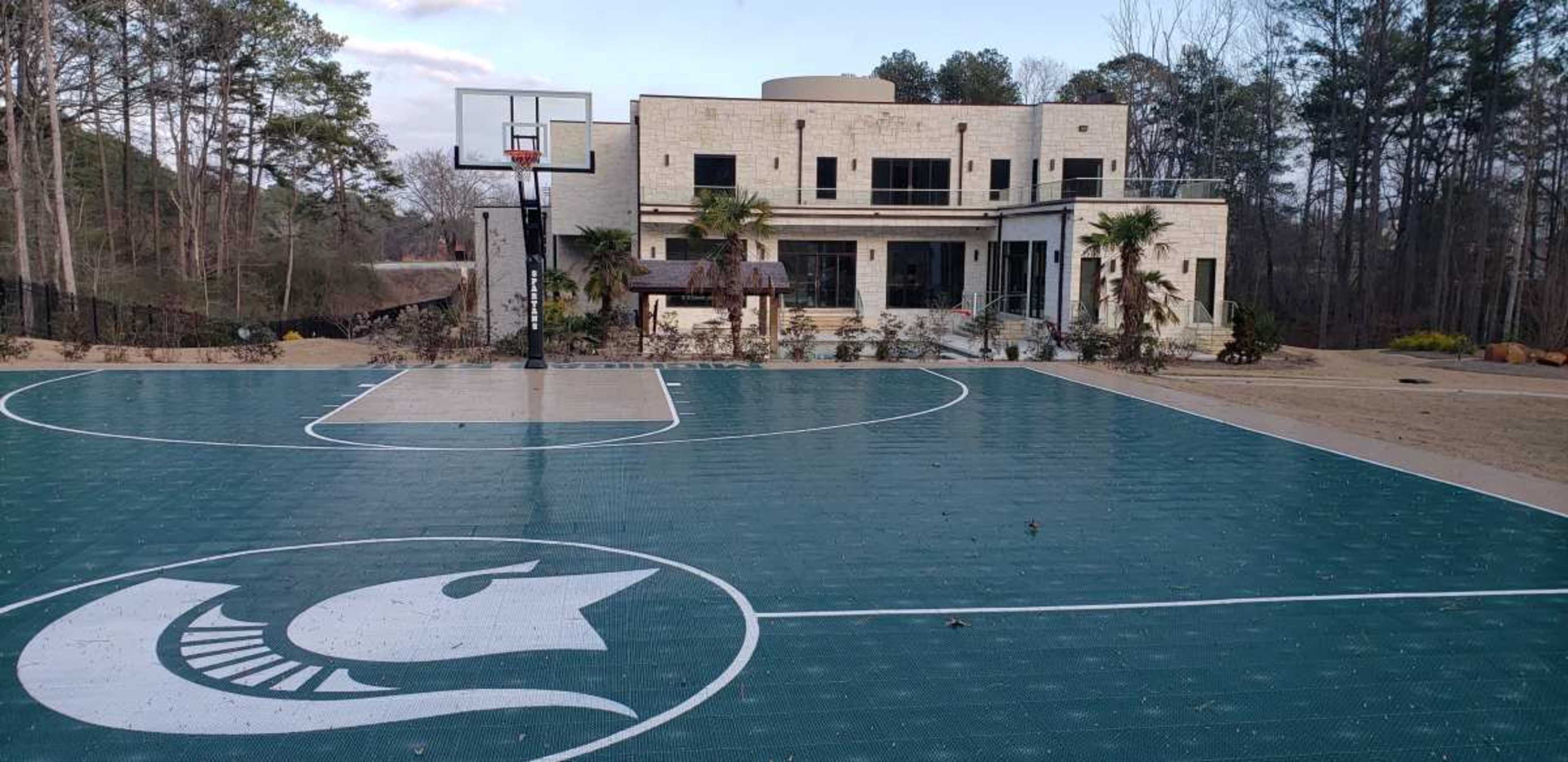A basketball court with a large logo in the center, situated in front of a modern house surrounded by trees.