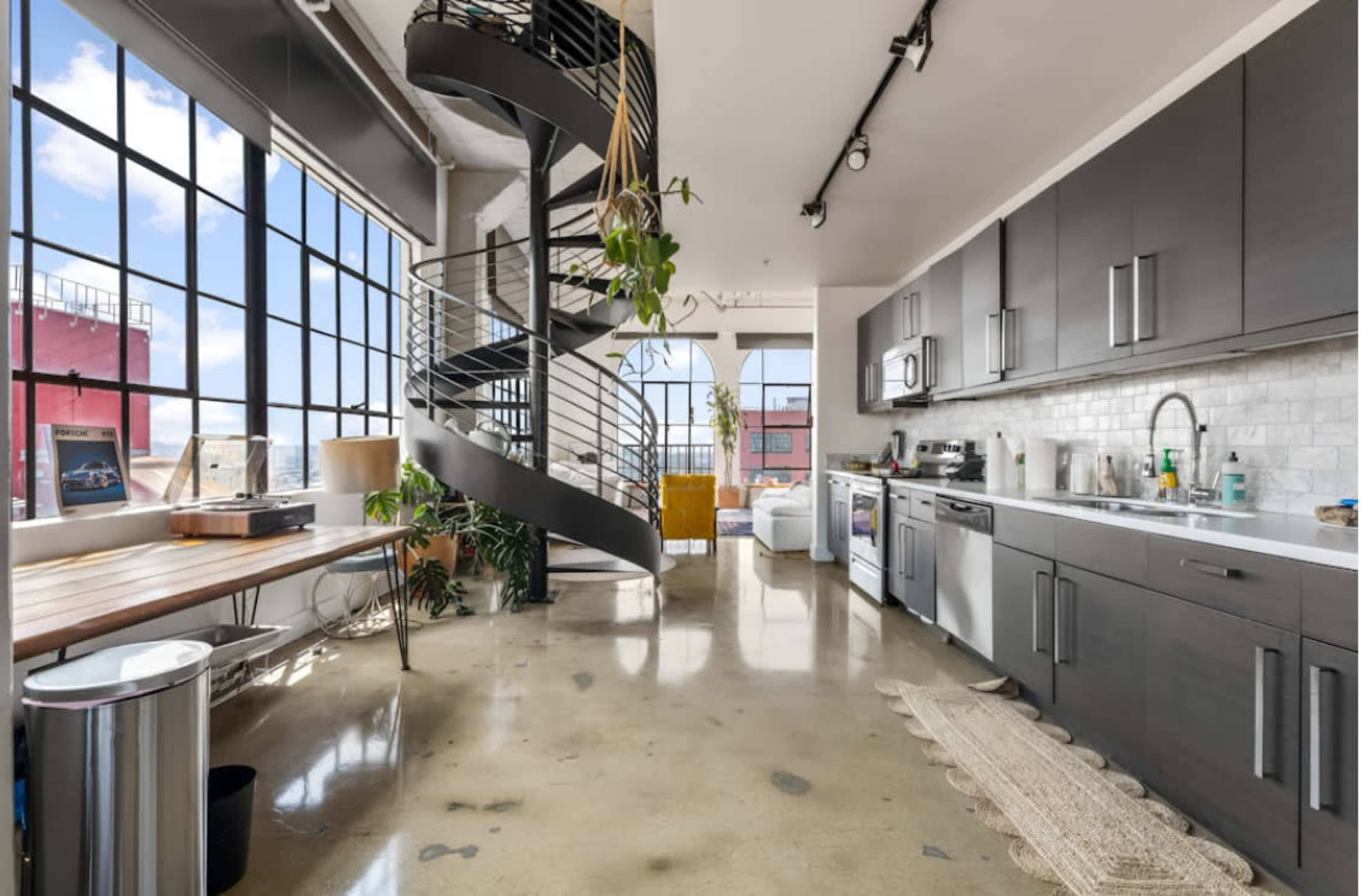 The image shows a modern kitchen and living area with a spiral staircase, large windows letting in natural light, and a polished concrete floor.