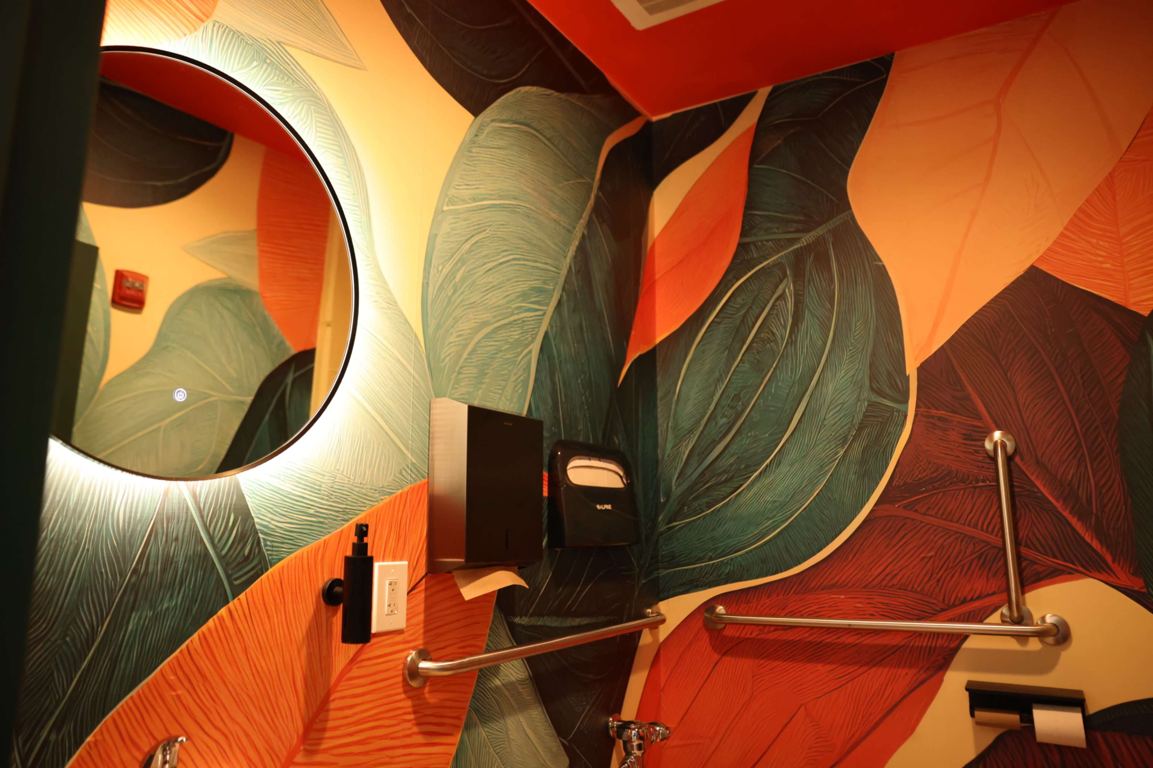 The image shows a bathroom with a colorful leaf-patterned mural on the walls, a round mirror, and a paper towel dispenser mounted beside a grab bar.