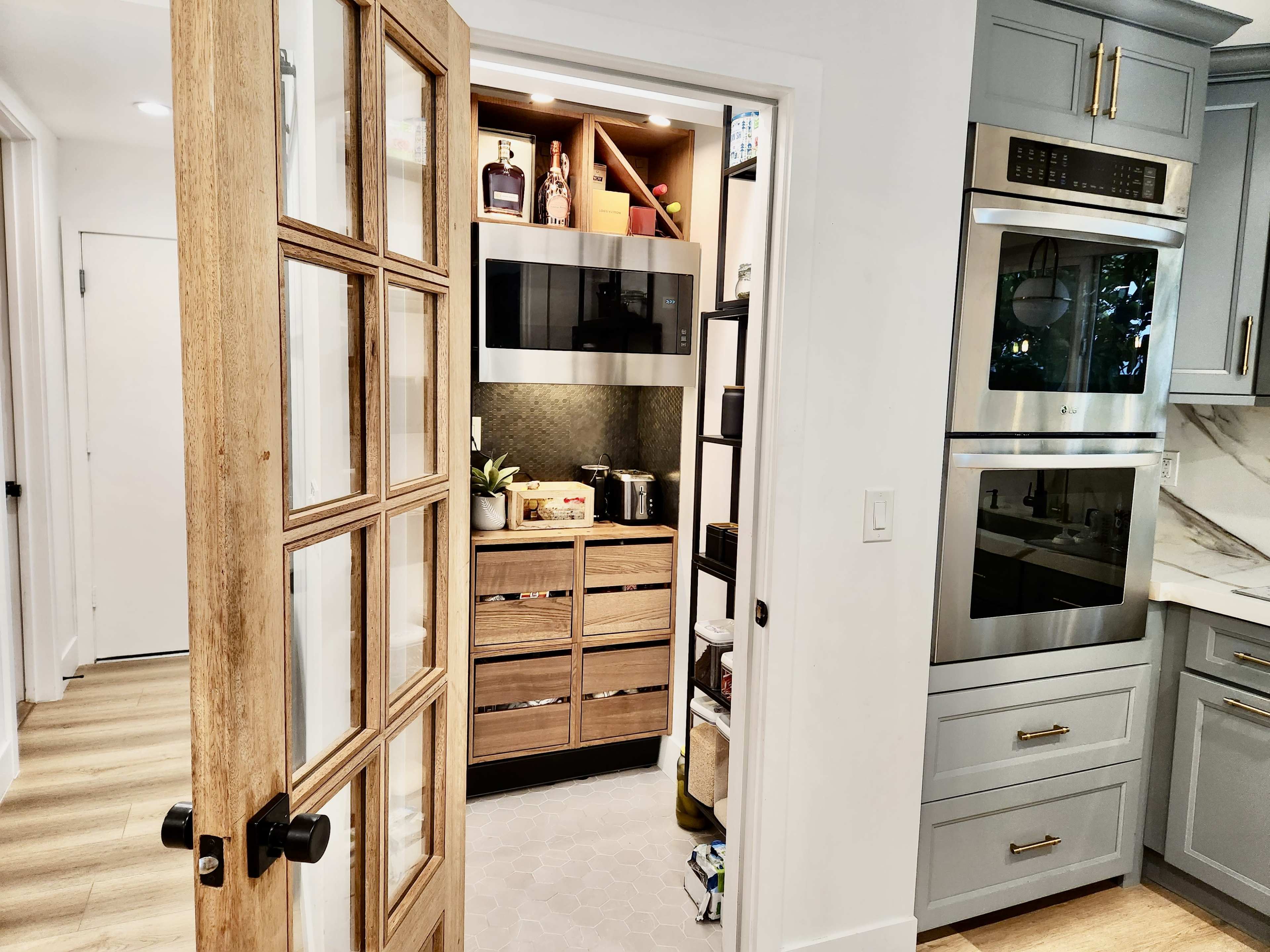 A wooden door opens to reveal a pantry area with organized shelves, a microwave, and several kitchen appliances.