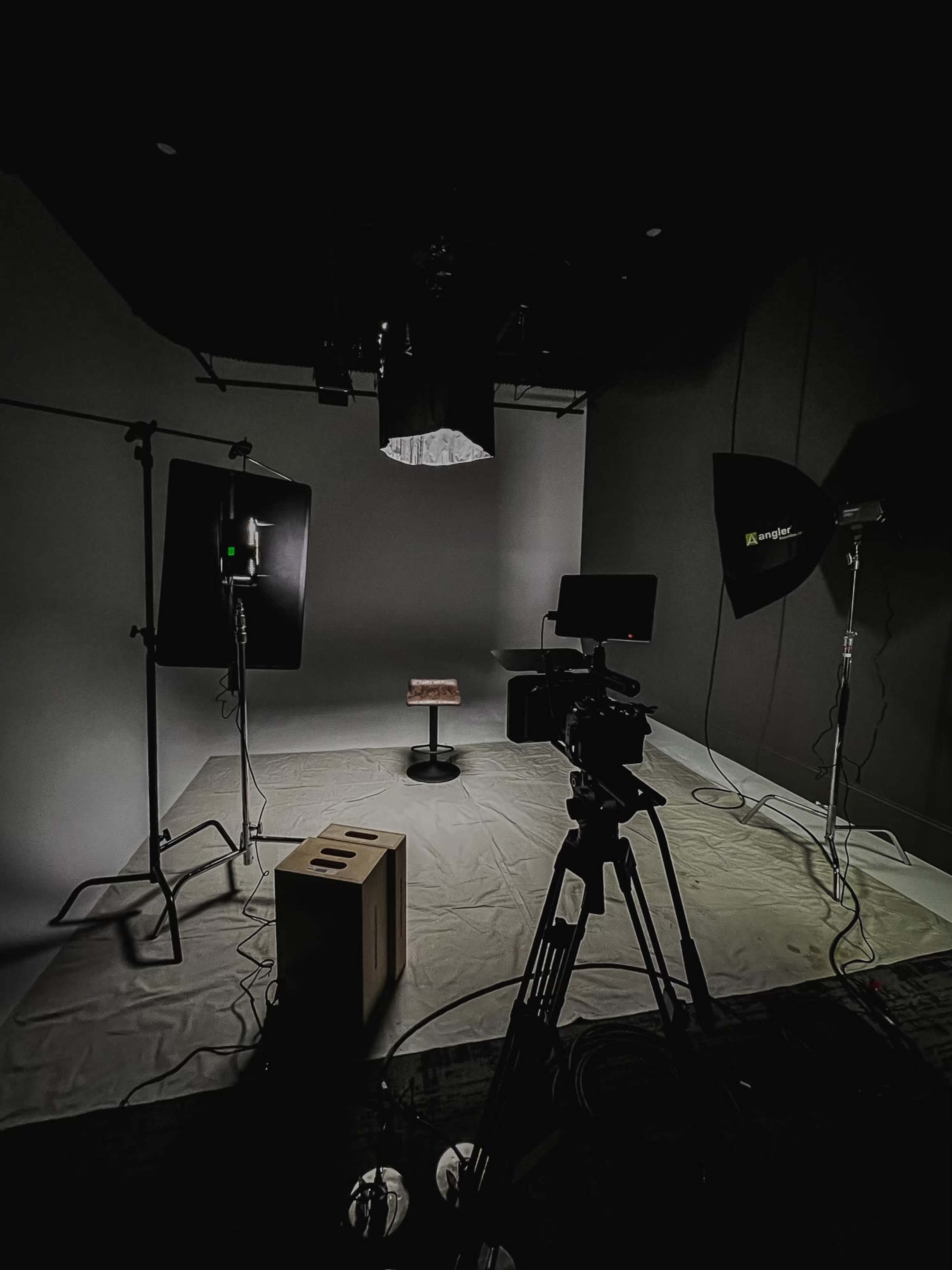 A dimly lit photography studio with a stool in the center, surrounded by lighting equipment and a video camera on a tripod.