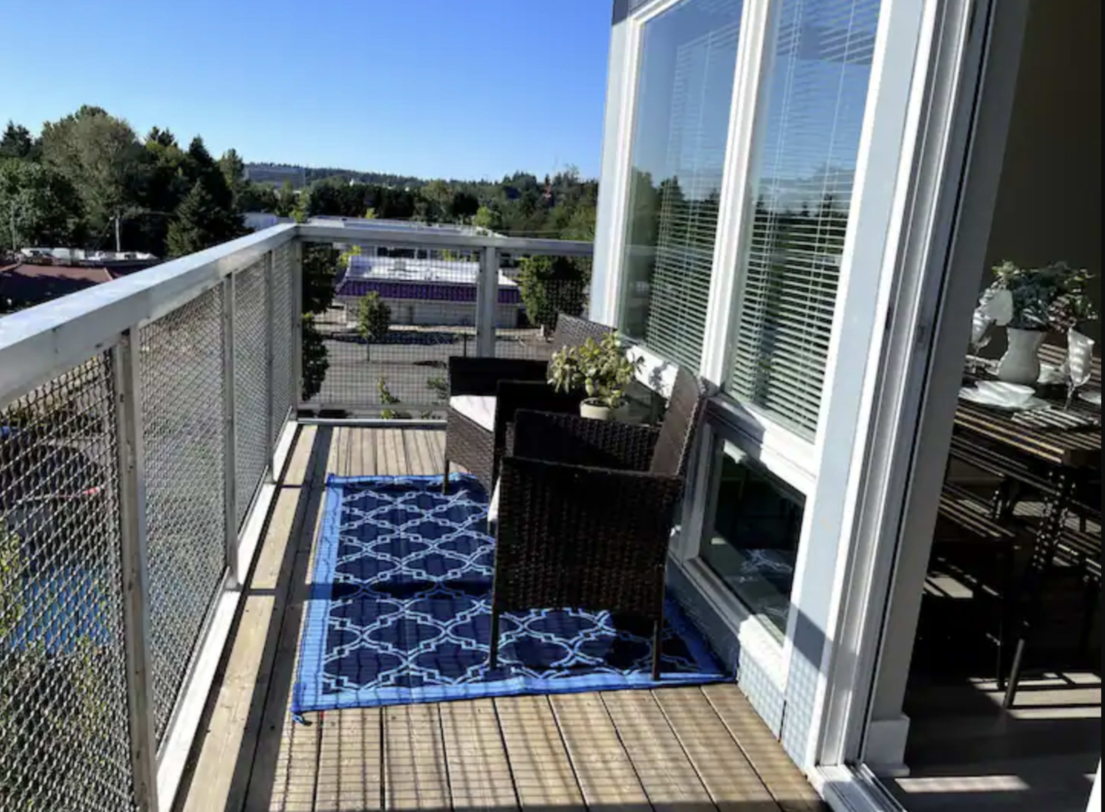 The image shows a balcony with two wicker chairs and a small potted plant on a blue patterned rug, overlooking a view of trees and a distant landscape.