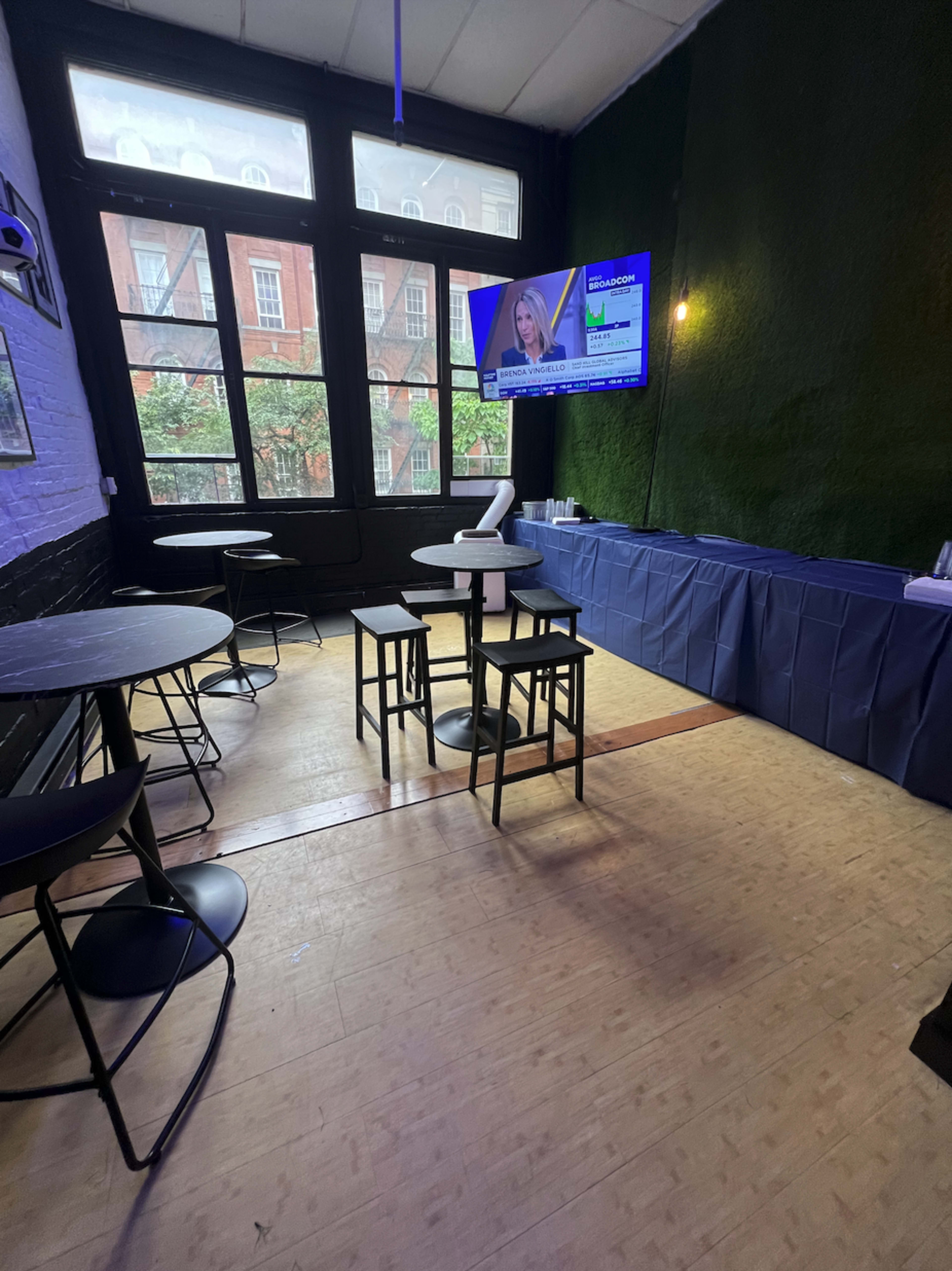 The image shows a modern indoor space with several small tables and stools, a bar area covered with a blue tablecloth, and a large television displaying news in front of large windows.