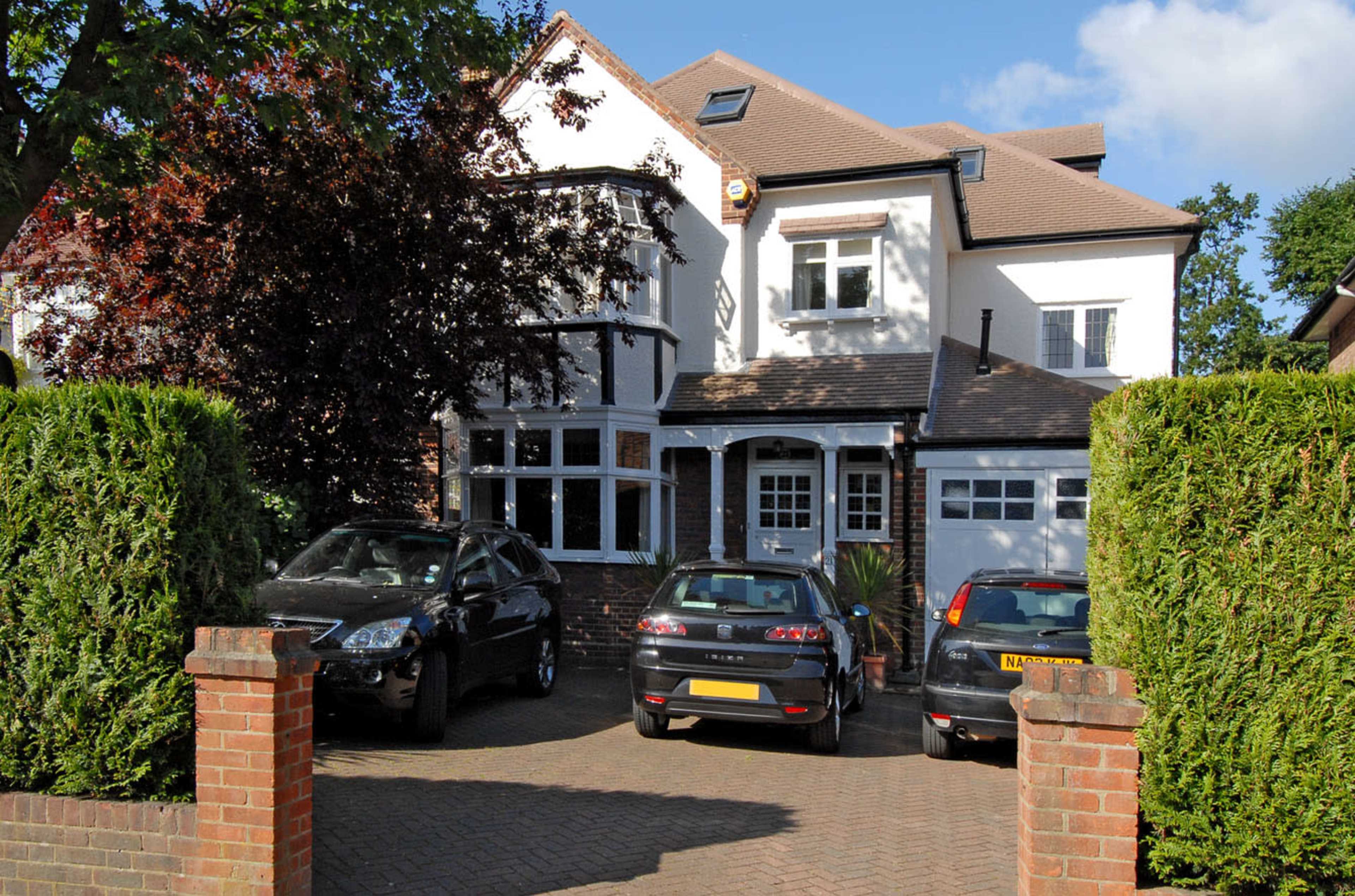 A two-story house with a front driveway contains four parked cars and is flanked by hedges.