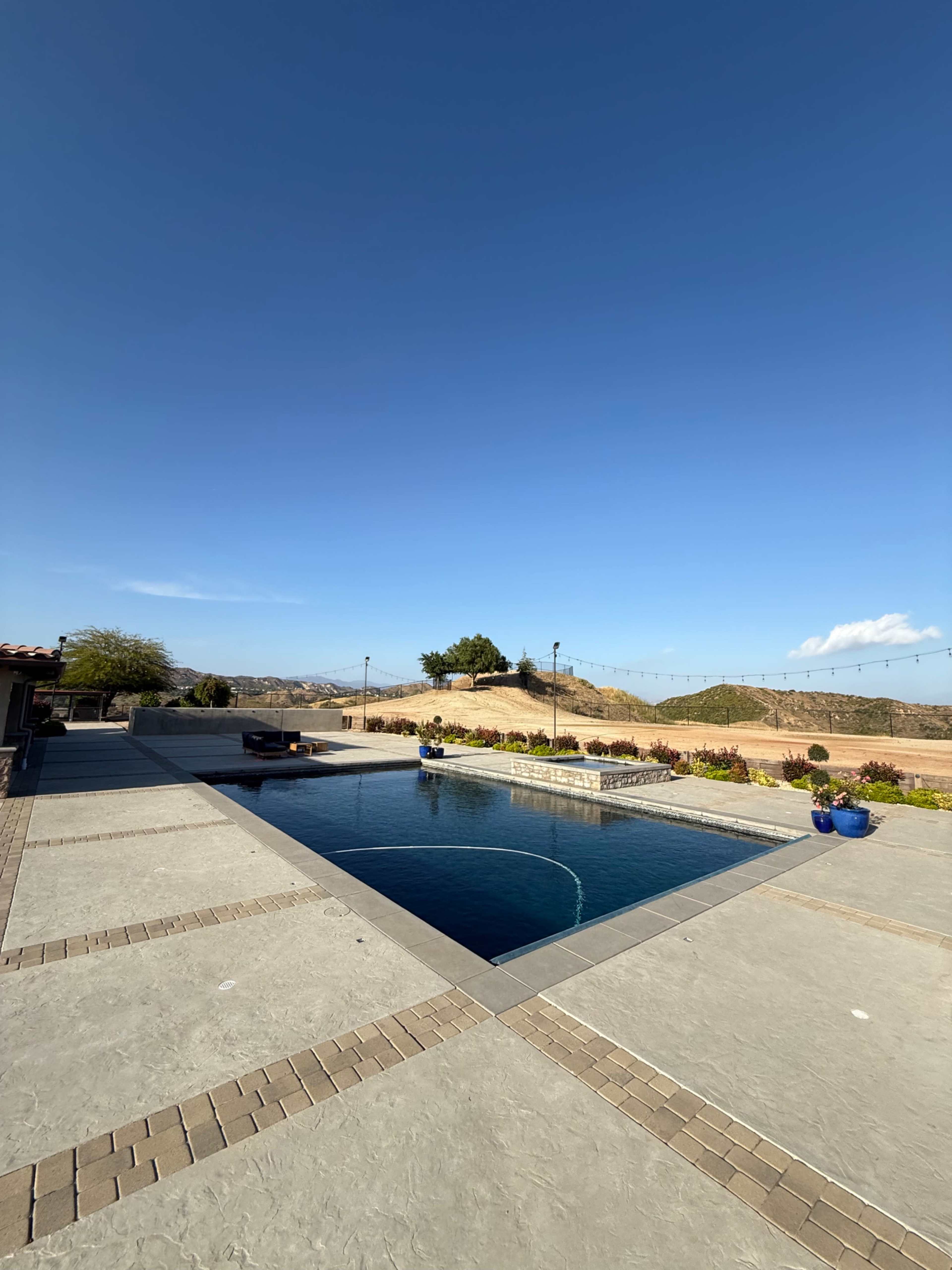 The image shows a swimming pool surrounded by a concrete deck, with a hillside and clear blue sky in the background.