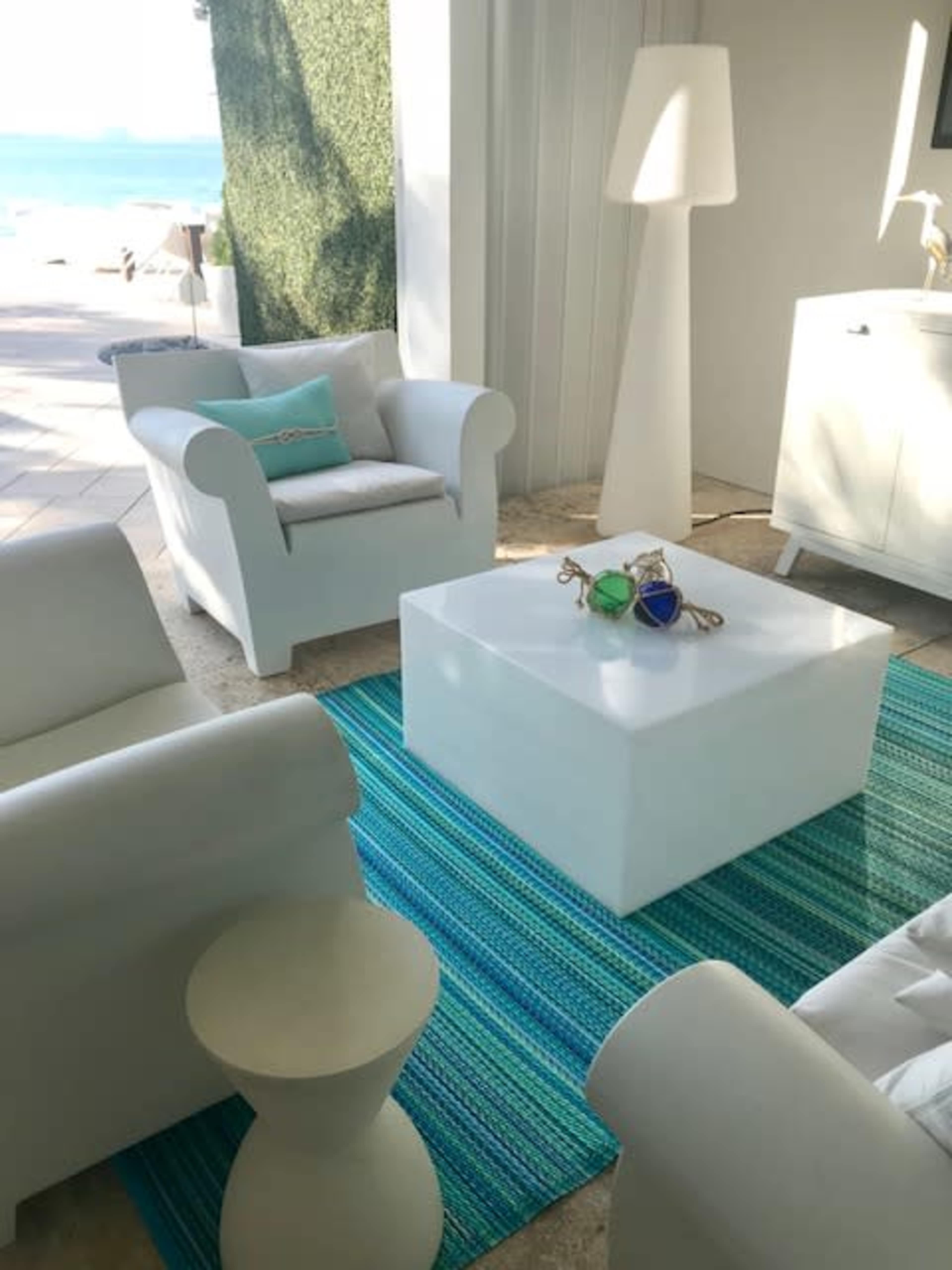 A modern lounge area features white furniture, including two chairs and a low table, on a blue and green striped rug, with a view of the ocean in the background.