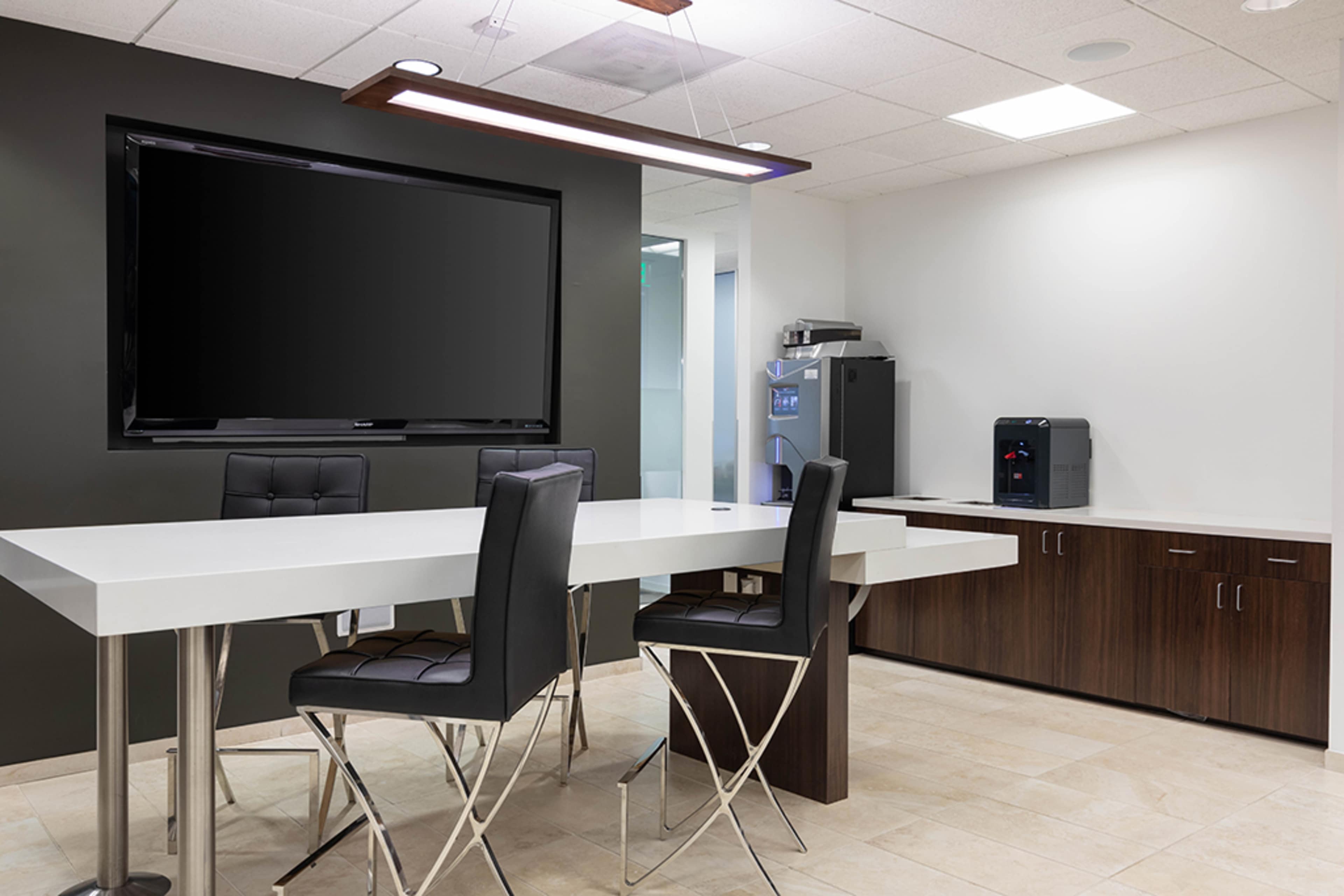 The image shows a modern meeting room with a large flat-screen television, a white table surrounded by black chairs, and wooden cabinetry along one wall.