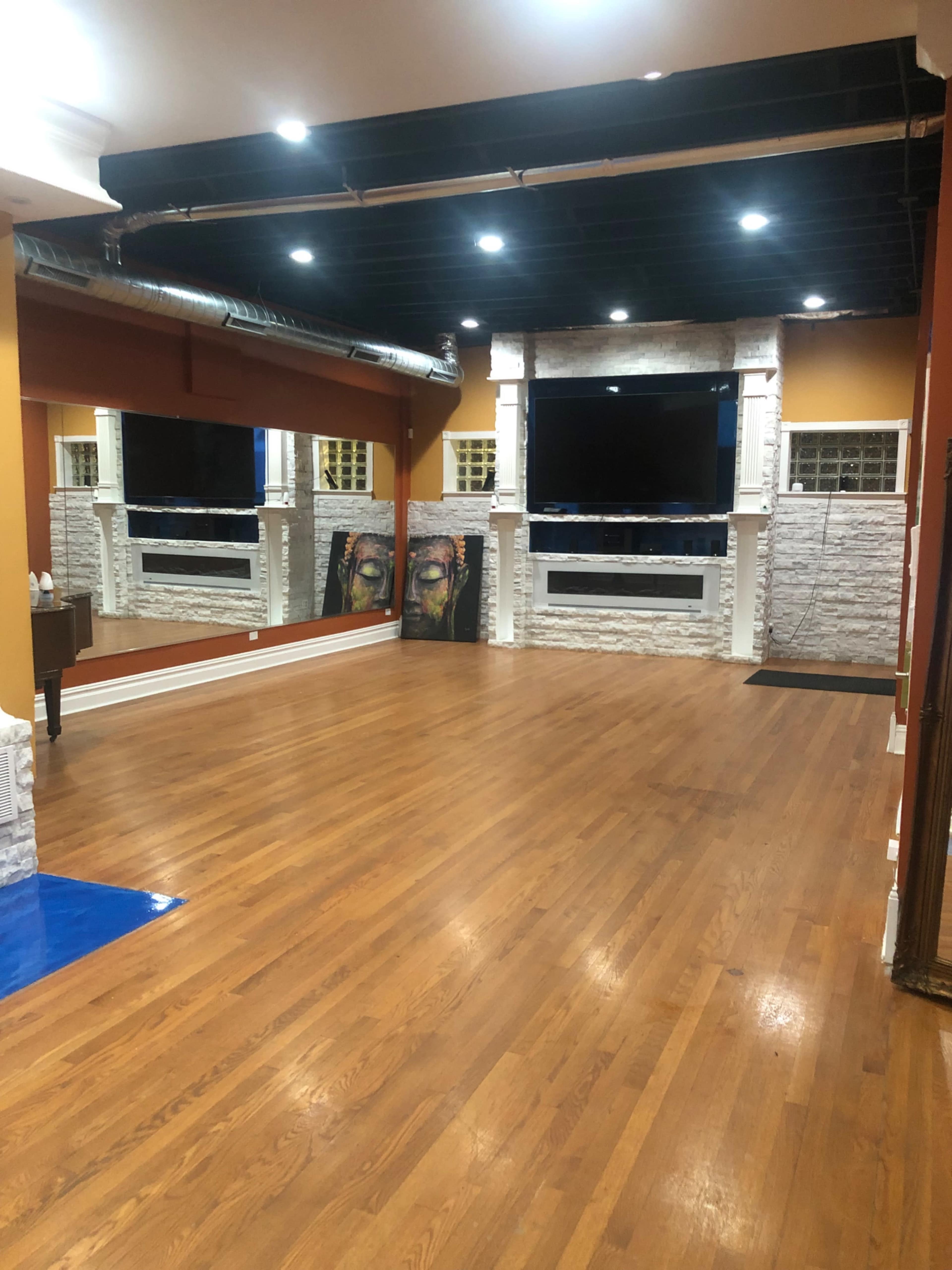 A spacious room with polished wooden floors, featuring a large flat-screen TV mounted above a stone fireplace, flanked by two framed artworks on the walls.