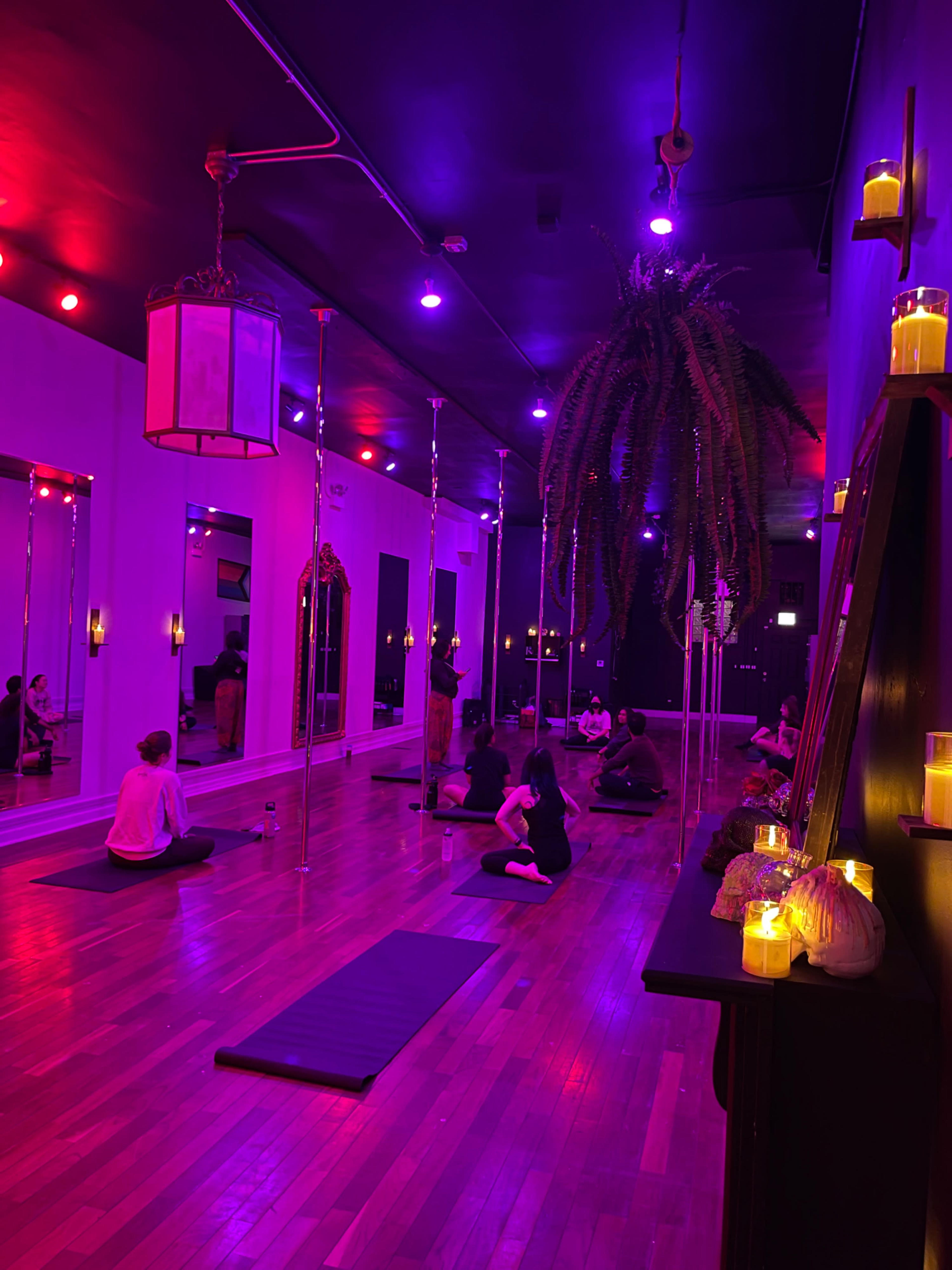 A dimly lit yoga studio with a wooden floor, featuring several people sitting on mats and surrounded by colorful lights and candles.