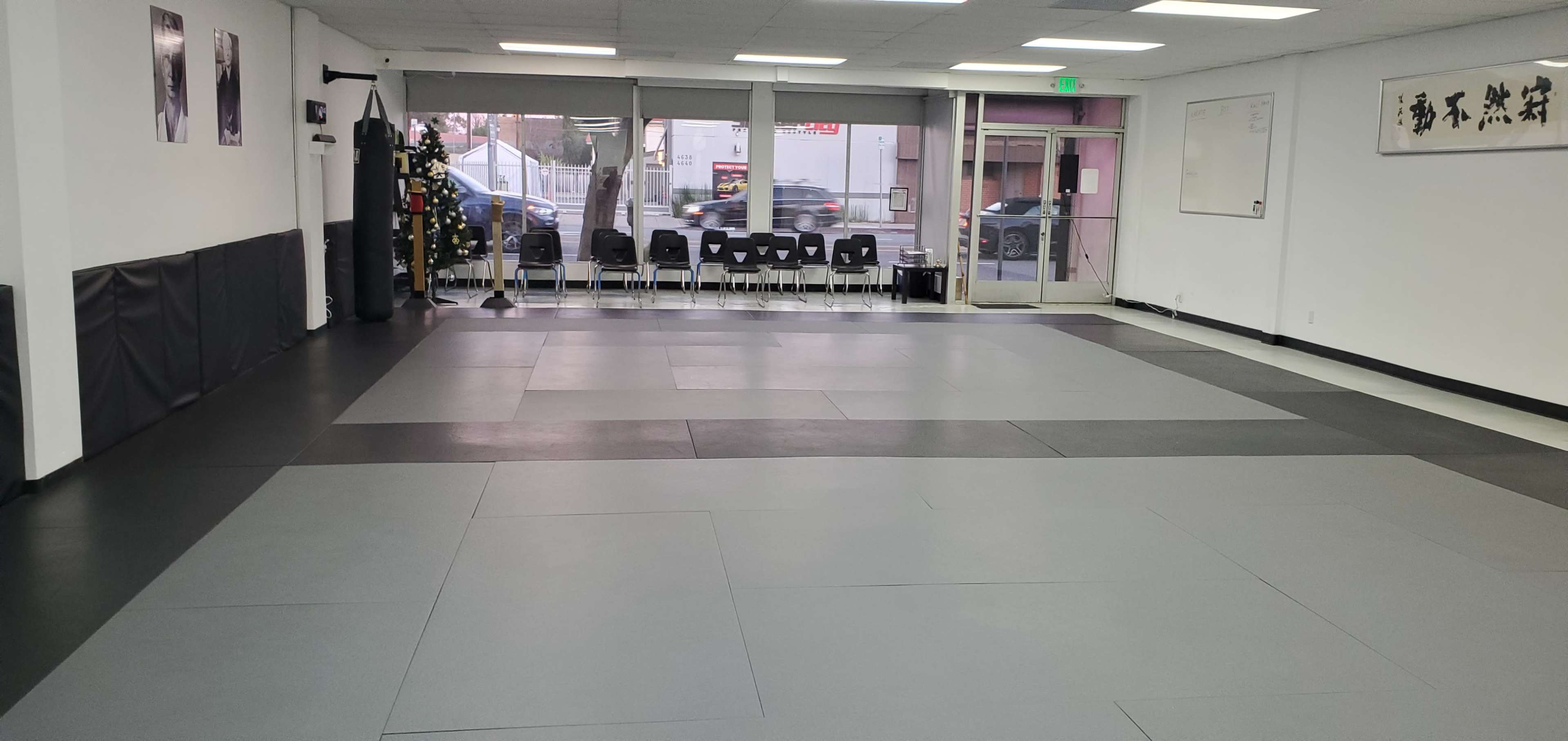 A spacious martial arts studio with gray mats, several chairs lined against a wall, and large front windows.