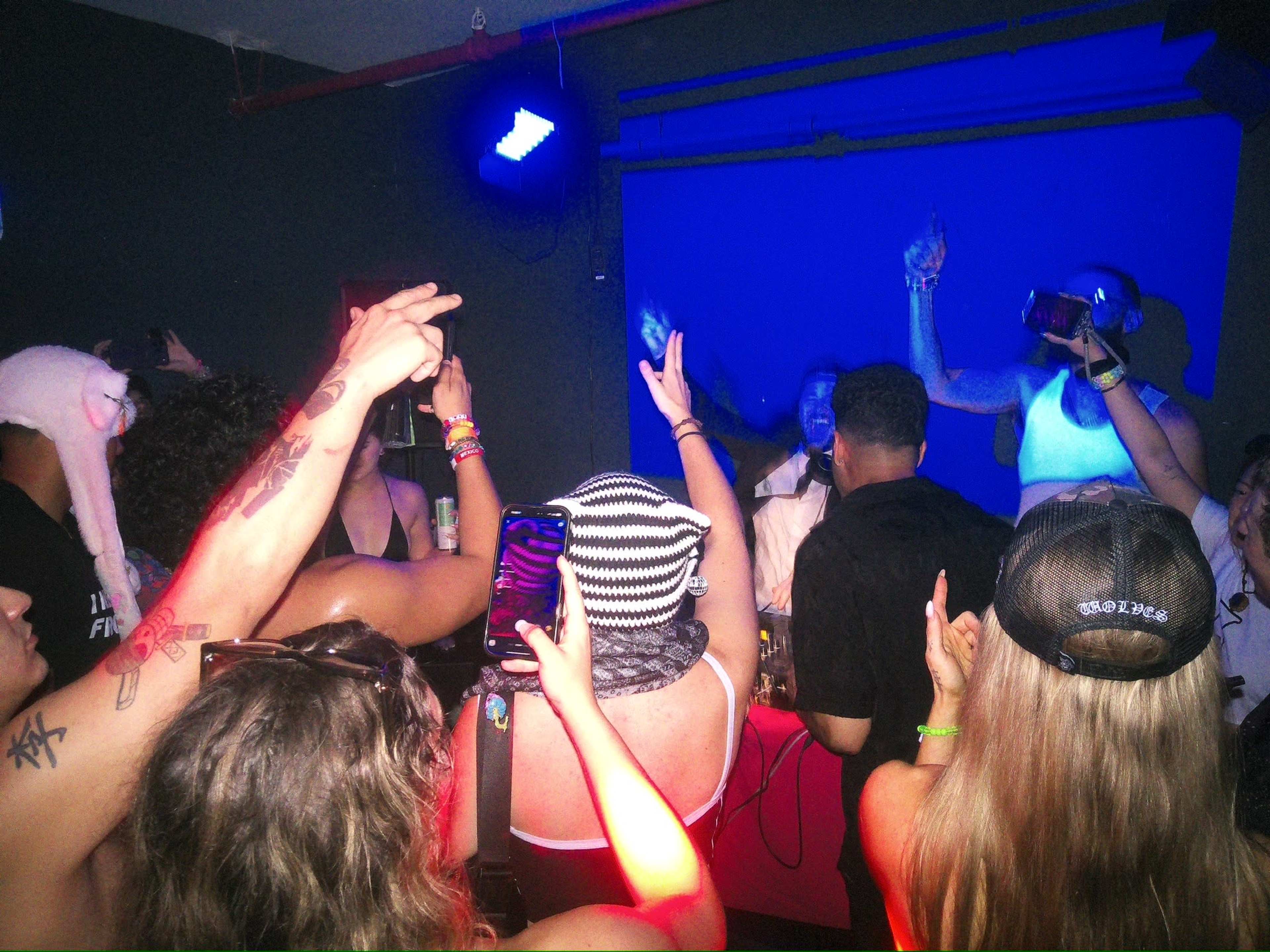 A group of people with their arms raised is gathered around a DJ at a lively indoor party, surrounded by colorful lights.
