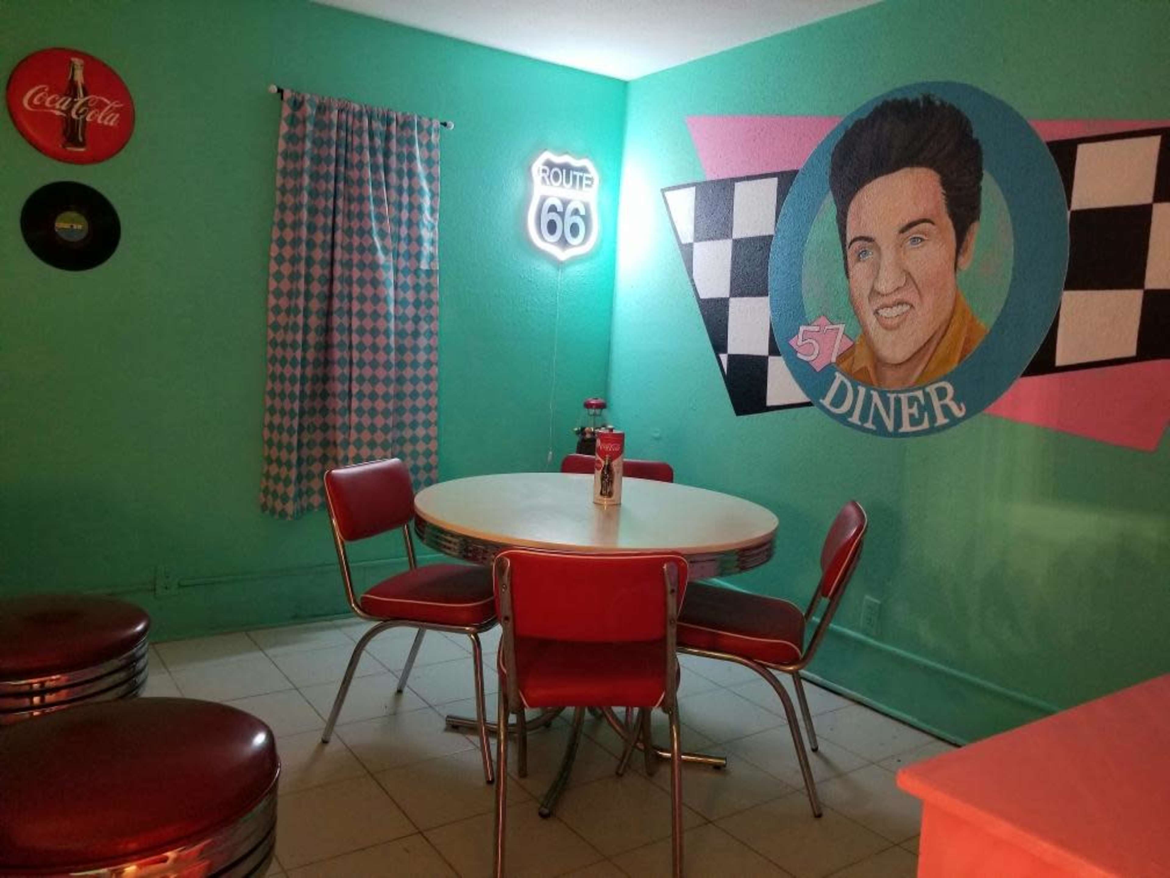 A retro diner setting with a round table and red chairs, a mural of a man resembling Elvis, and a blue-green wall decorated with Route 66 memorabilia.