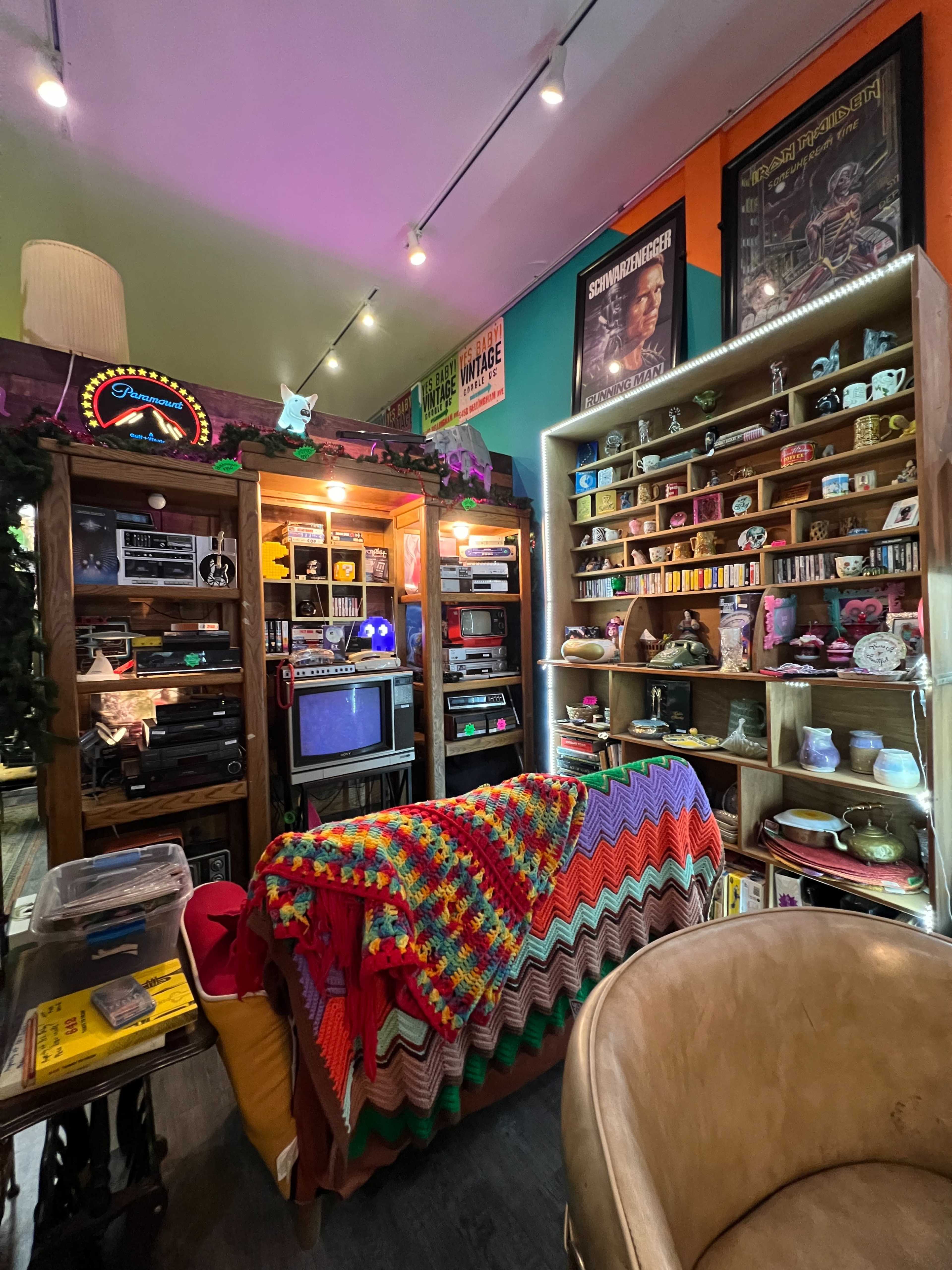 The scene features a cozy vintage shop interior filled with shelves displaying various collectibles, electronics, and colorful decor, alongside a crocheted blanket draped over a chair.