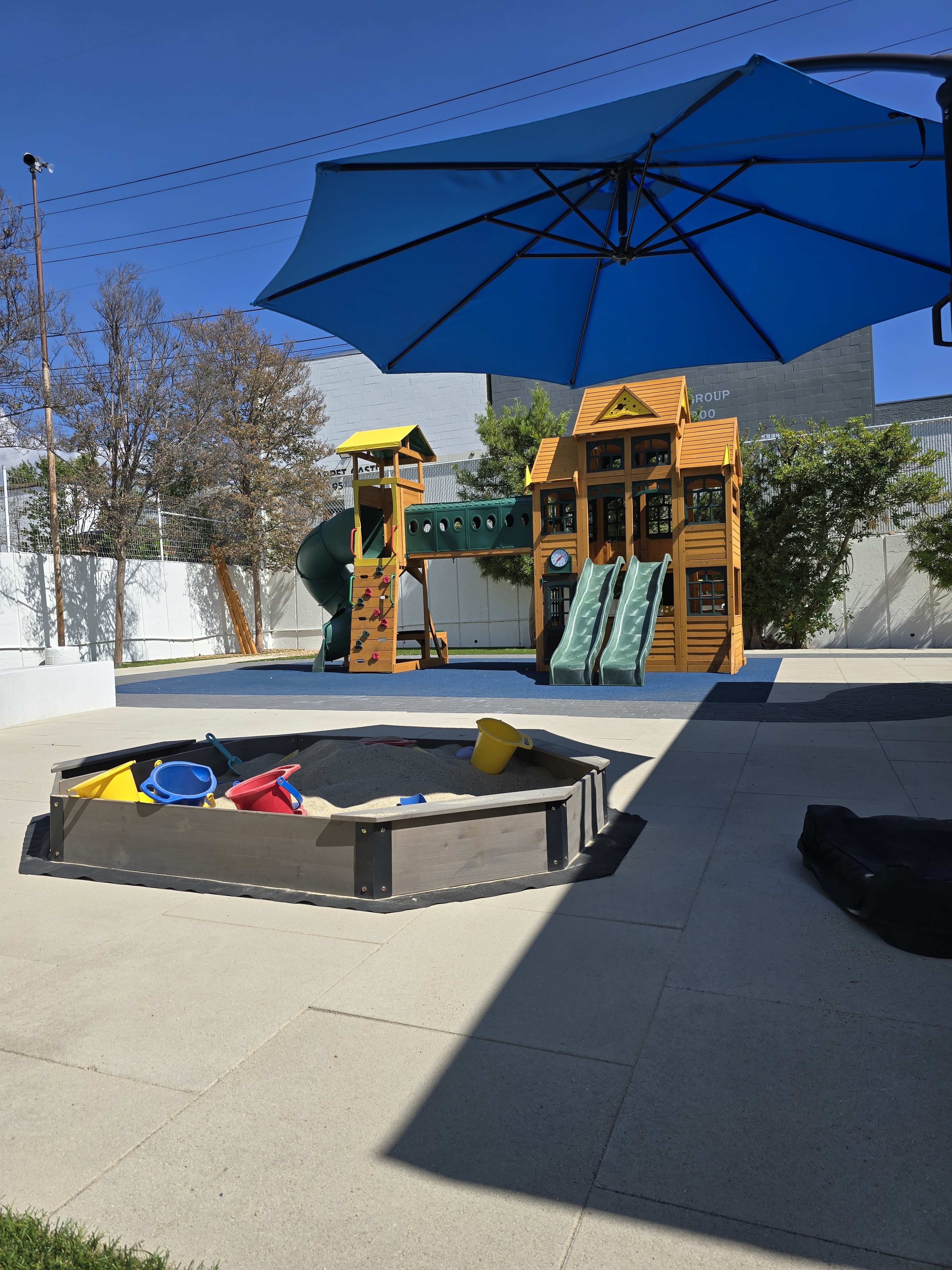 The image shows a colorful playground with a slide and climbing structures, alongside a sandpit equipped with various toys, all under a large blue umbrella.