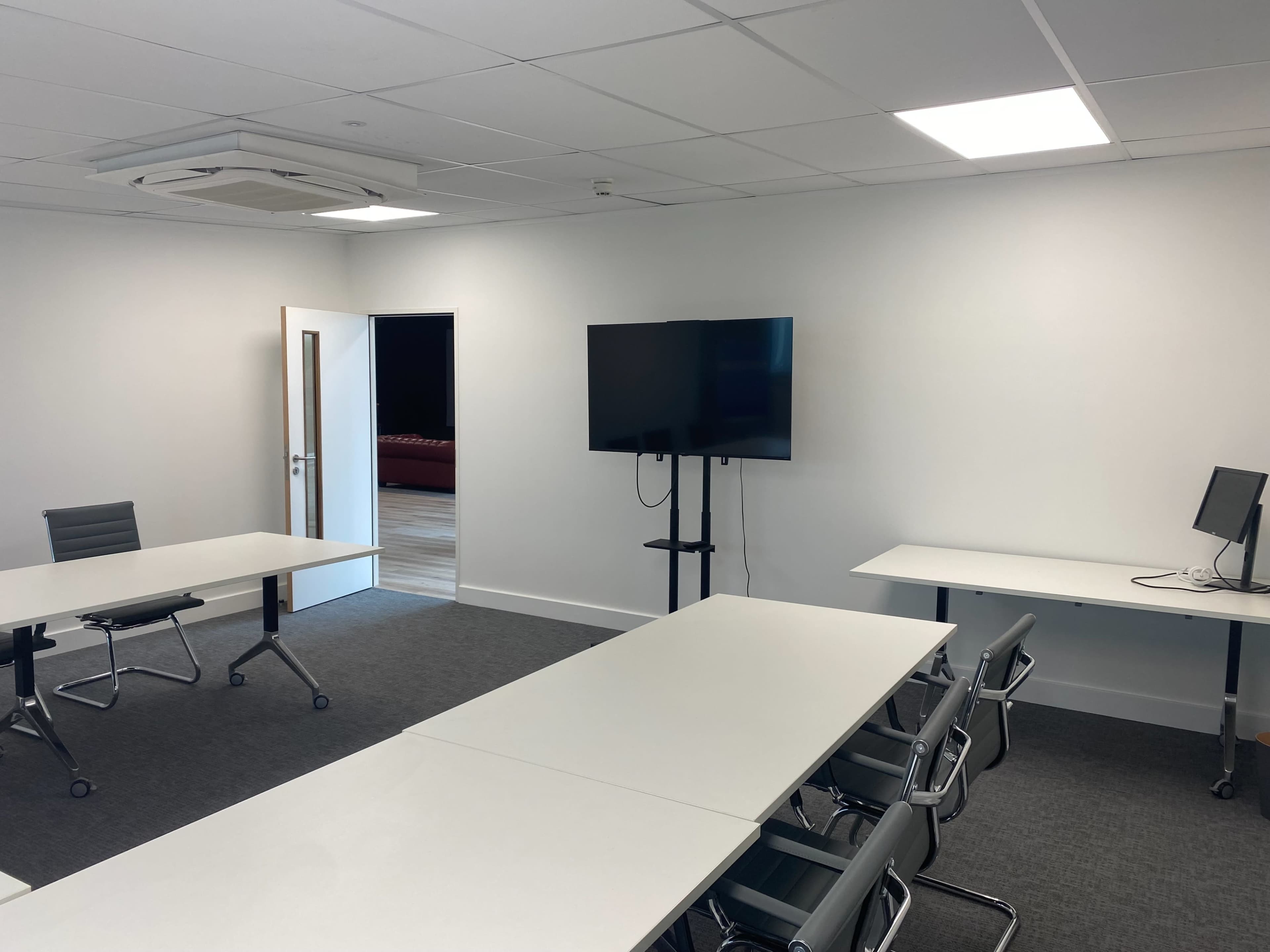 The image shows a modern conference room with two tables, a monitor on a stand, and a doorway leading to another room.
