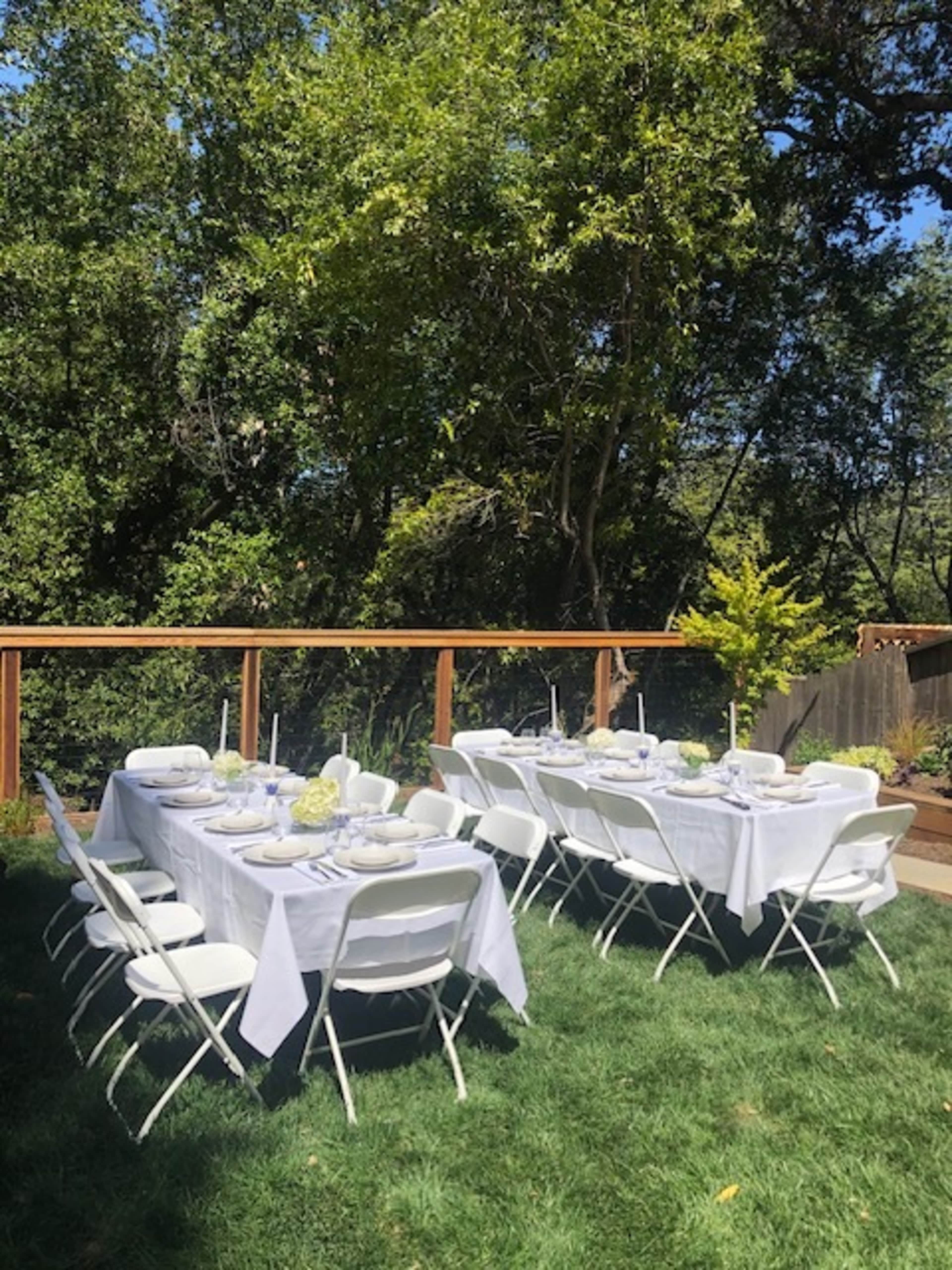 A long rectangular table is set with white tablecloths and dishes under a wooden deck surrounded by greenery.