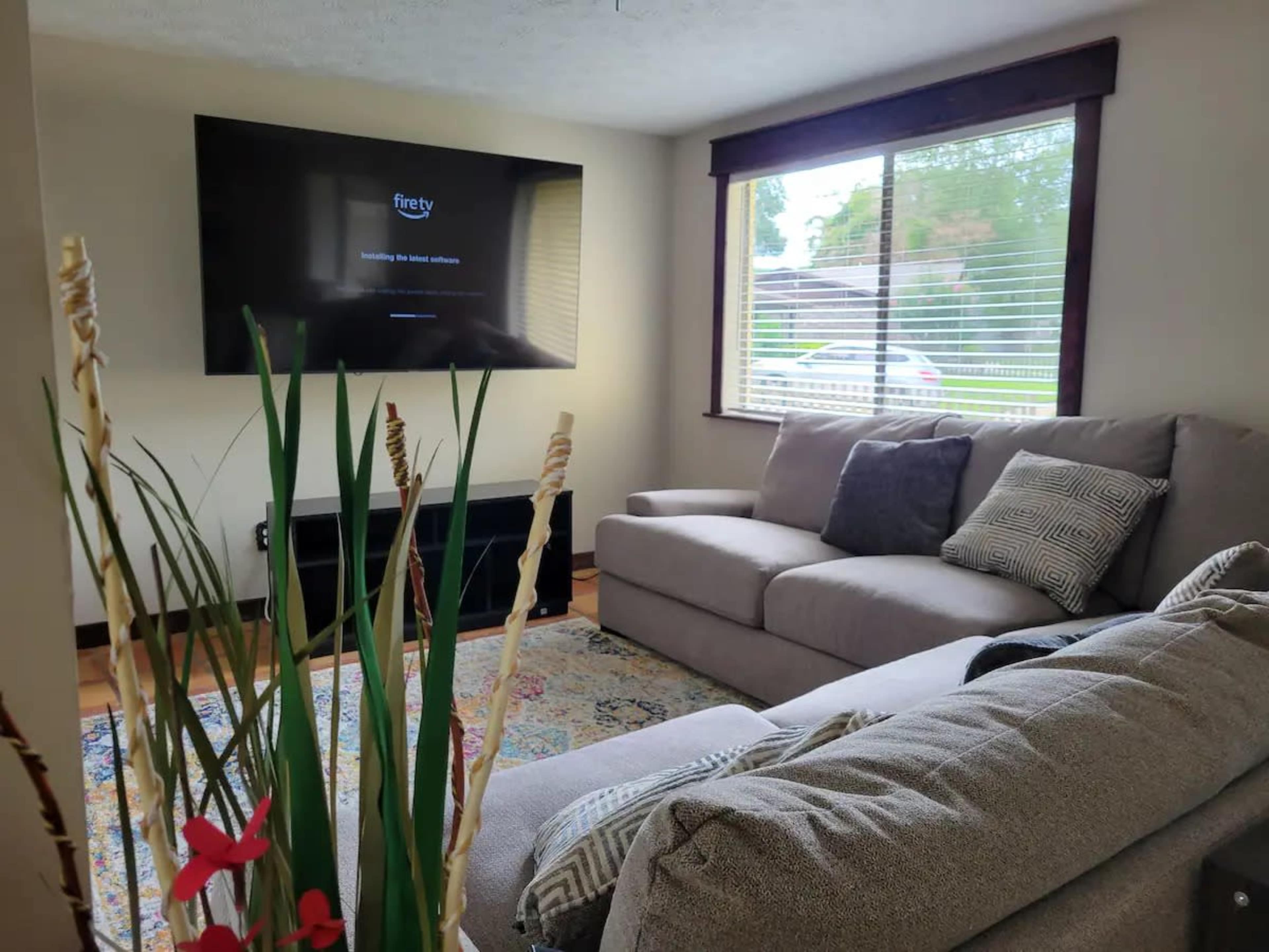 A living room with a sectional sofa, a television mounted on the wall, and a window with blinds letting in natural light.