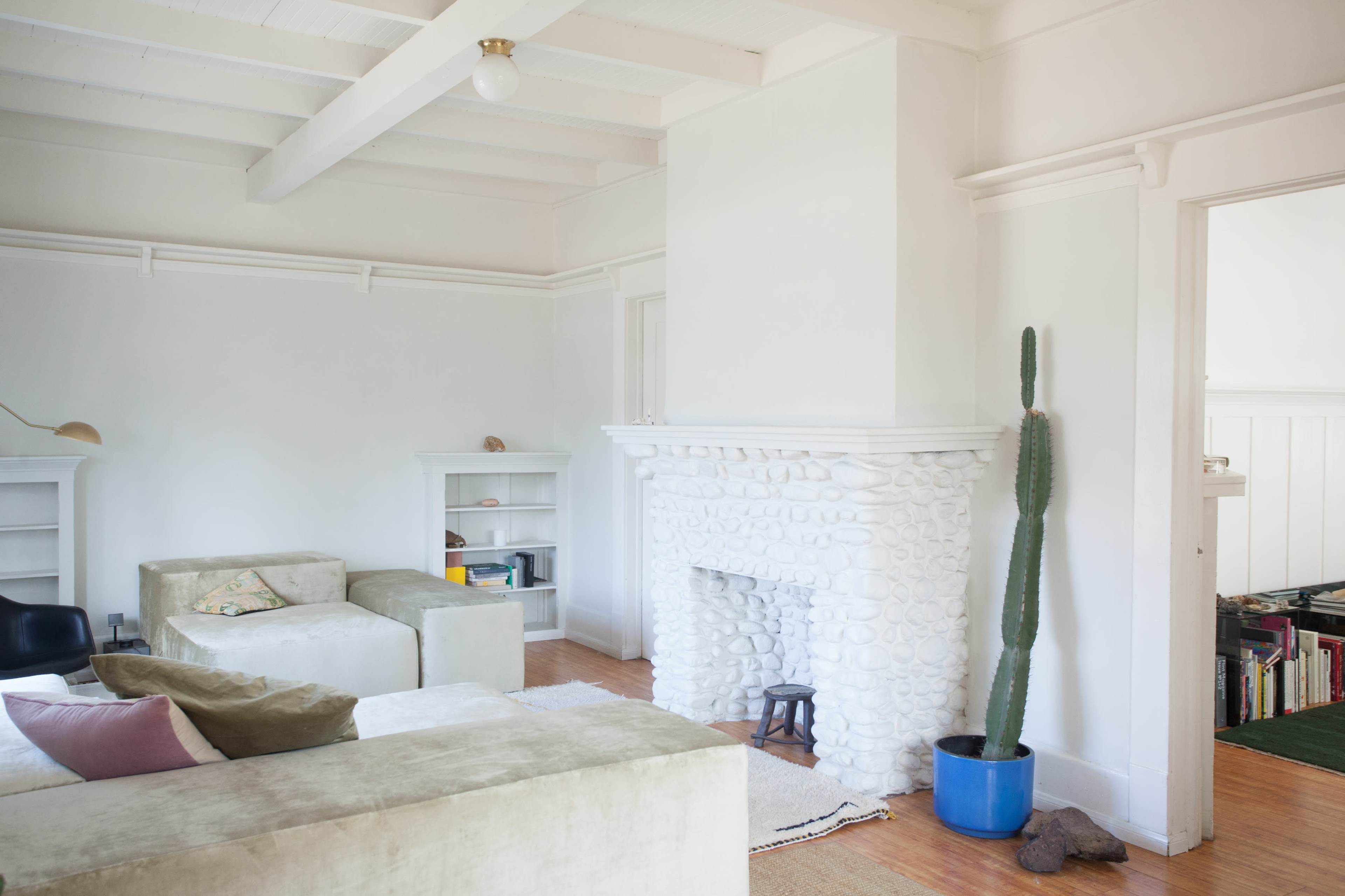 The image depicts a bright, minimalist living room featuring a white stone fireplace, a cactus in a pot, and a sectional sofa.