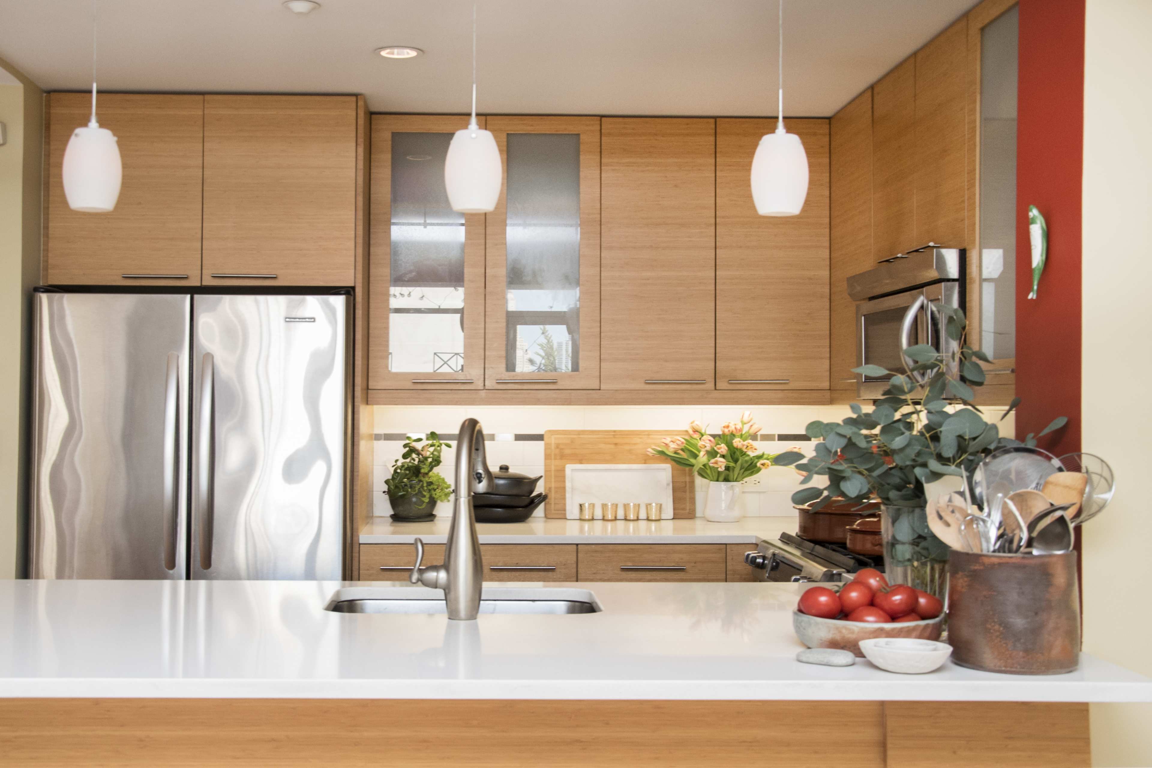 The image shows a modern kitchen with wooden cabinetry, stainless steel appliances, and a white countertop, featuring a bowl of red apples and various kitchen utensils.